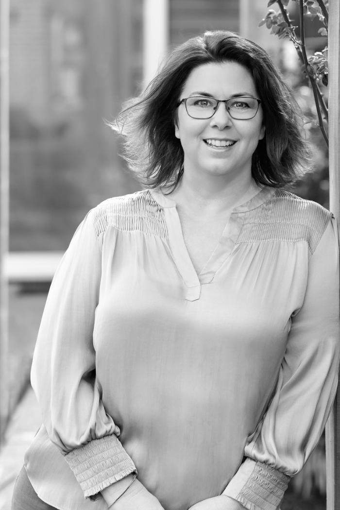 A woman with curly hair and glasses smiling outdoors, wearing a light-colored blouse with gathered cuffs.