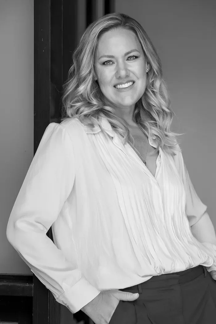 Black and white photo of a woman with wavy blonde hair, smiling, wearing a light-colored blouse and dark pants, standing indoors next to a wooden surface.