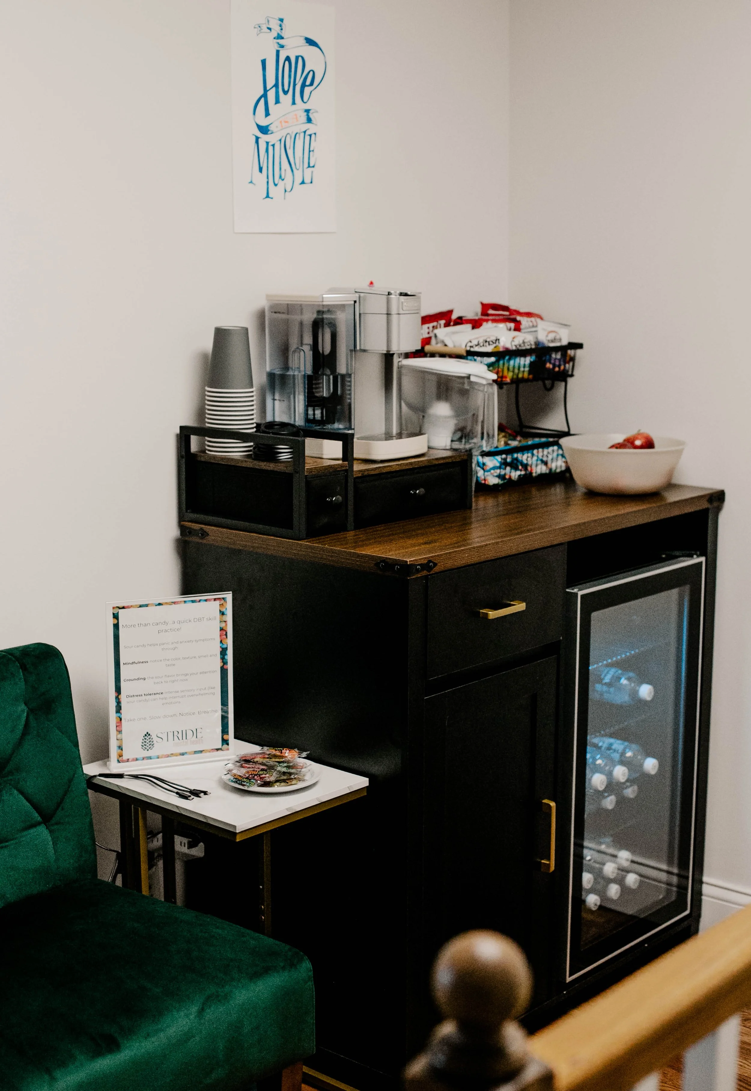 A small black bar cart with a coffee machine, cups, a bowl of apples, and snacks on top, next to a black wine fridge, a white side table with a sign, and a green velvet chair in a cozy corner of a room.