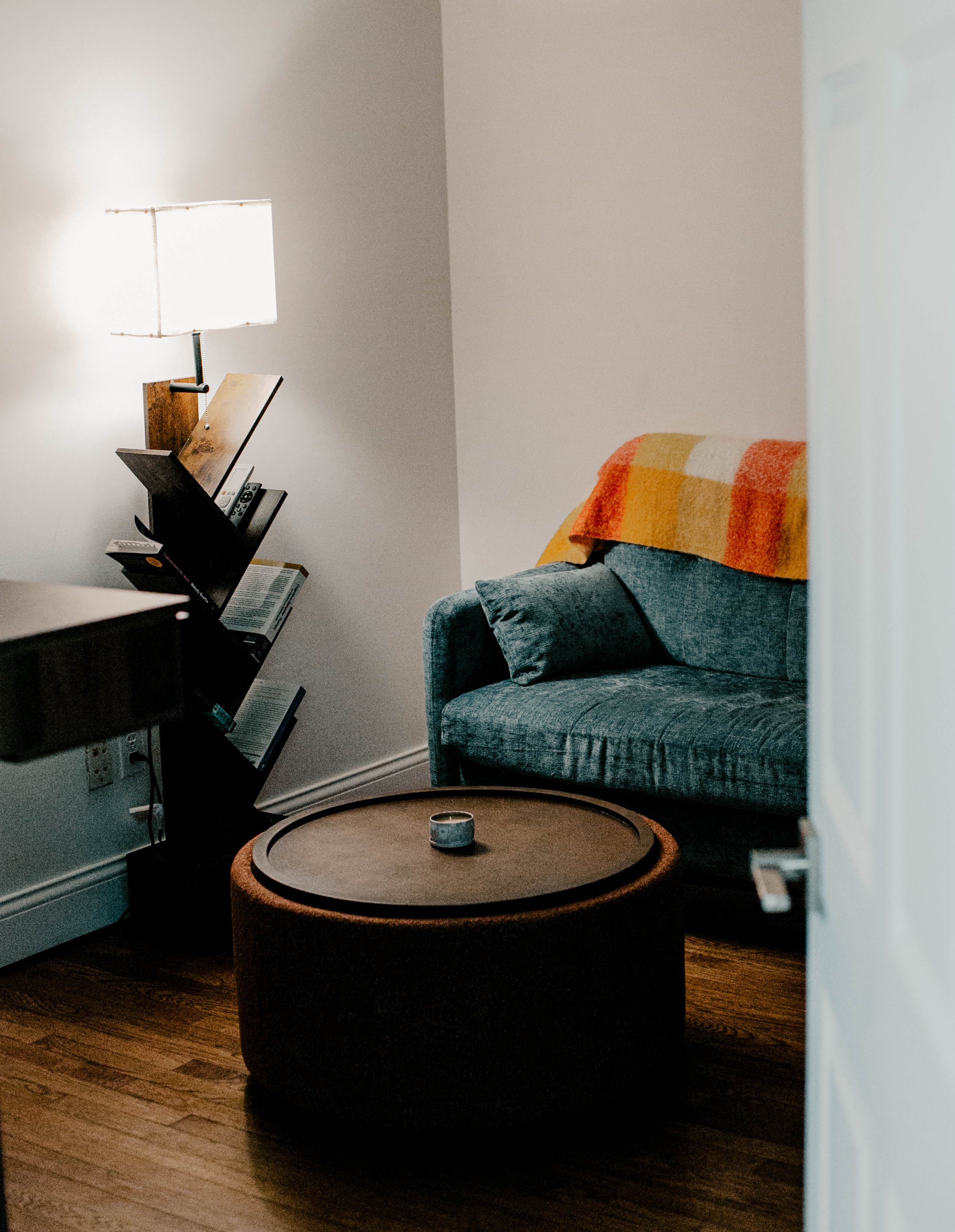 Cozy living room corner with a teal sofa covered with a striped orange and yellow blanket, a round ottoman, a bookshelf with books and remote controls, a table lamp, and hardwood floors.