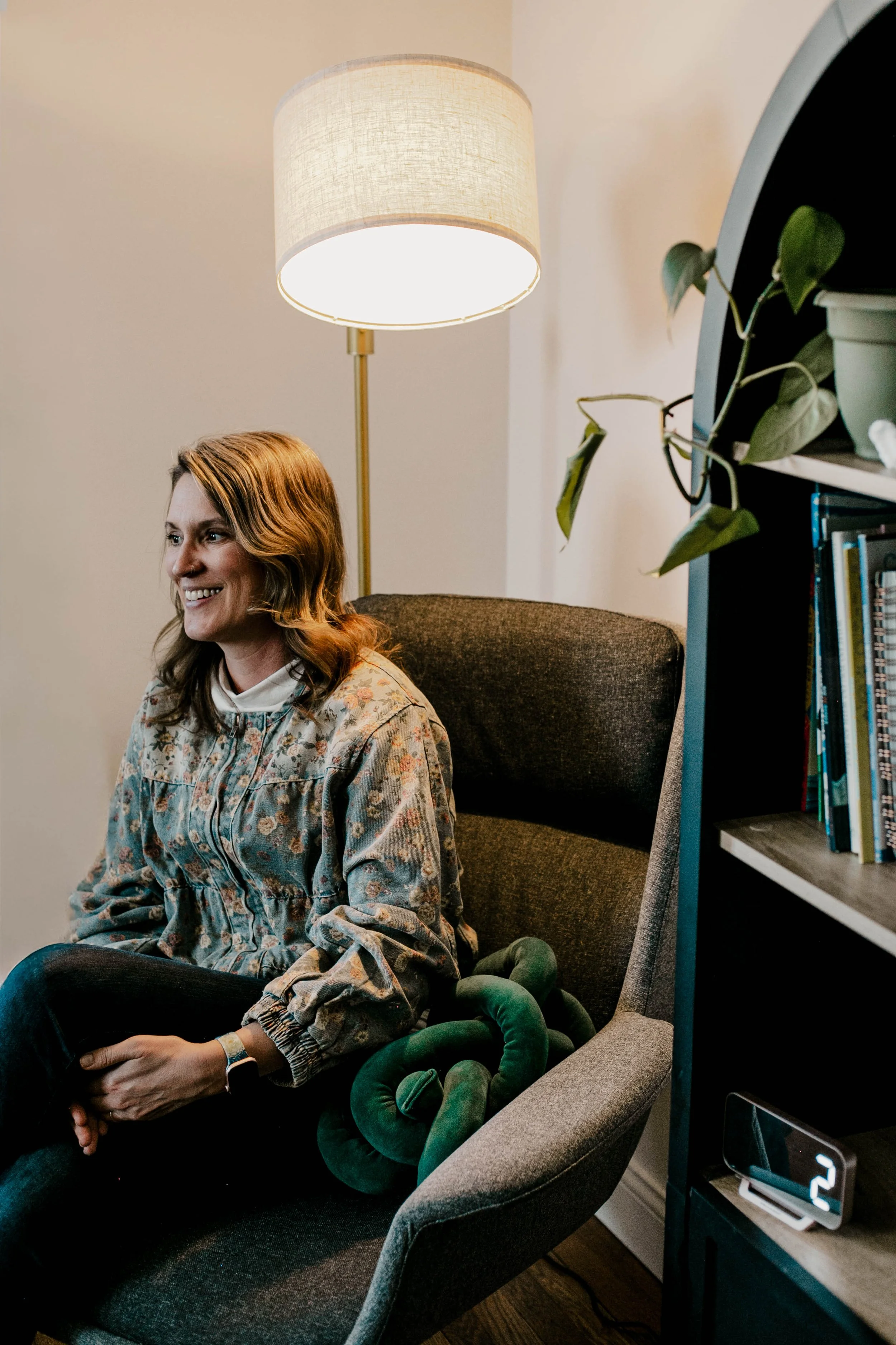 A woman with shoulder-length light brown hair smiling and sitting in a dark gray armchair, wearing a floral patterned shirt. Behind her, a tall floor lamp with a beige lampshade provides light. To her right, there's a black bookshelf with potted plants and books. A small digital clock showing the number 2 is on the shelf.