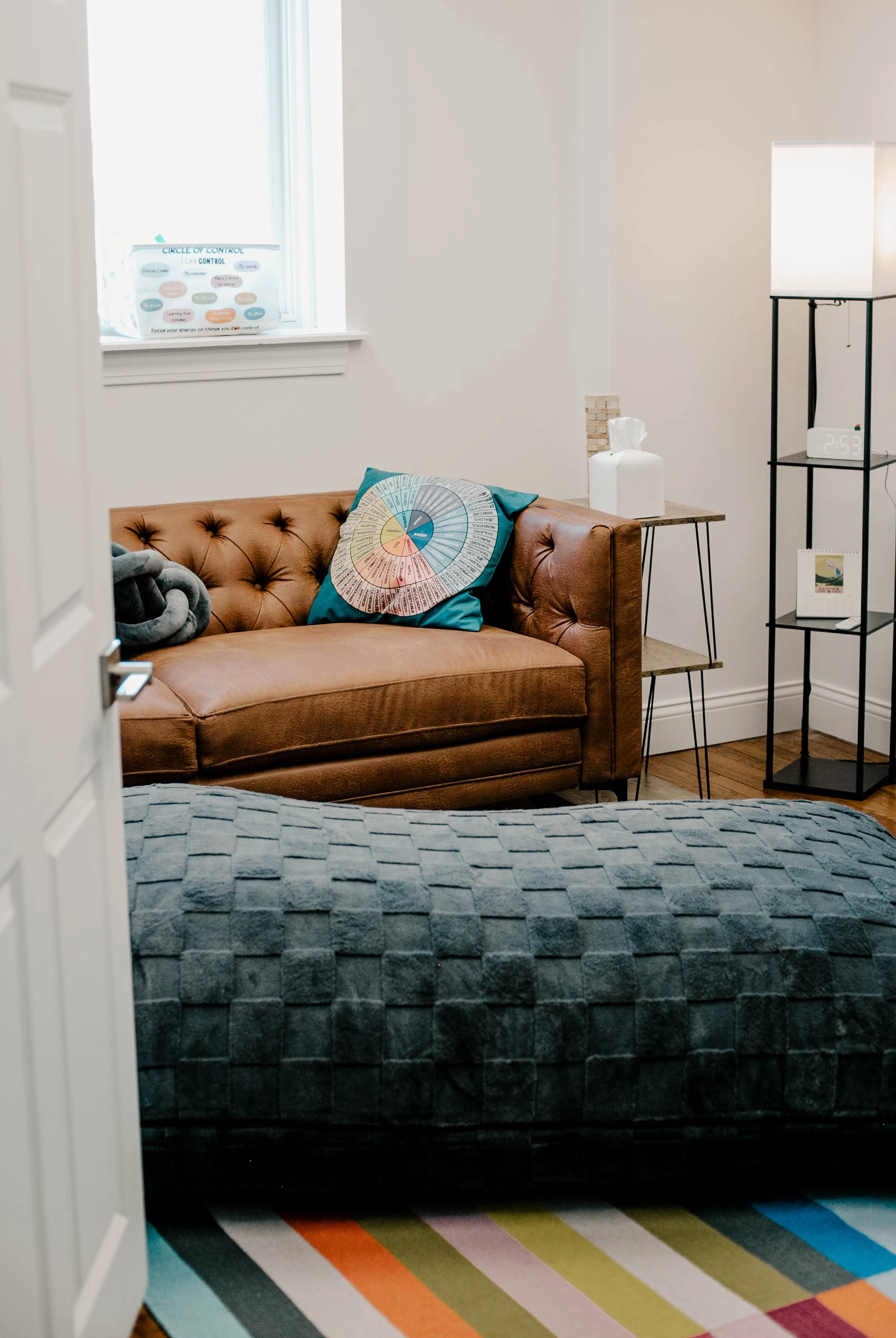 An interior scene of a cozy living room with a brown leather sofa, a dark gray textured blanket on the bed in the foreground, a colorful circular pillow on the sofa, a wooden side table with a white tissue box, a black metal shelf with a digital clock, a framed picture, a small lamp, and a window with natural light.