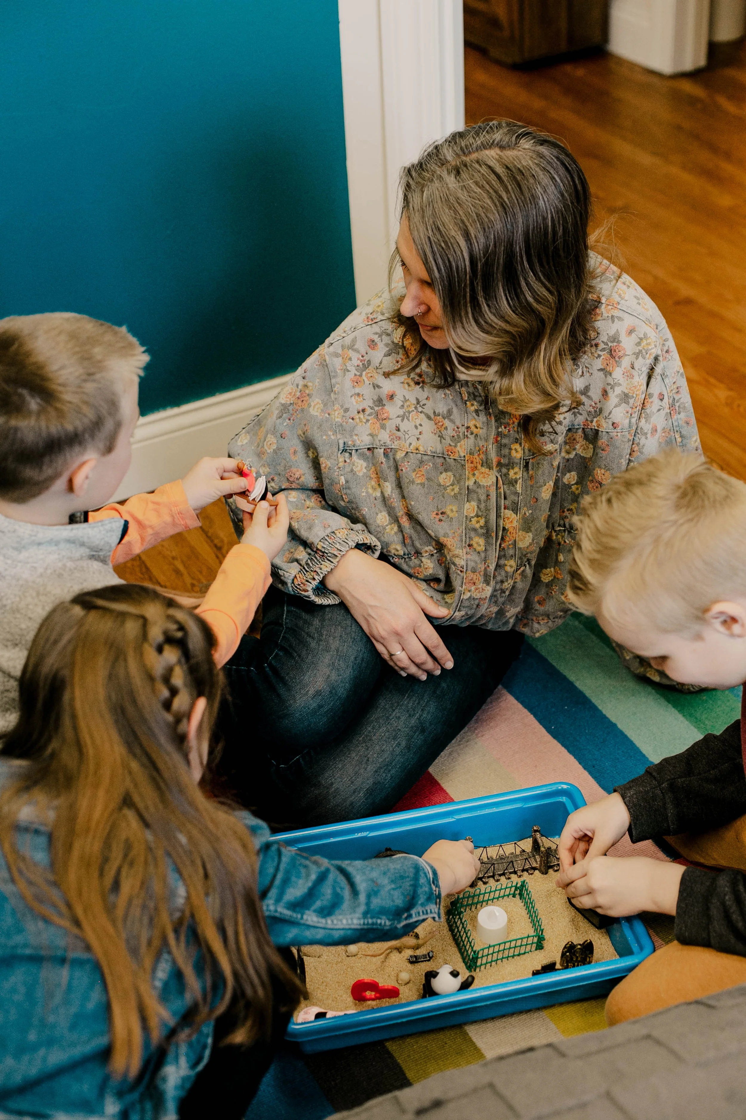 A woman sitting on the floor with four children around a plastic container filled with small toys, engaged in a play activity.