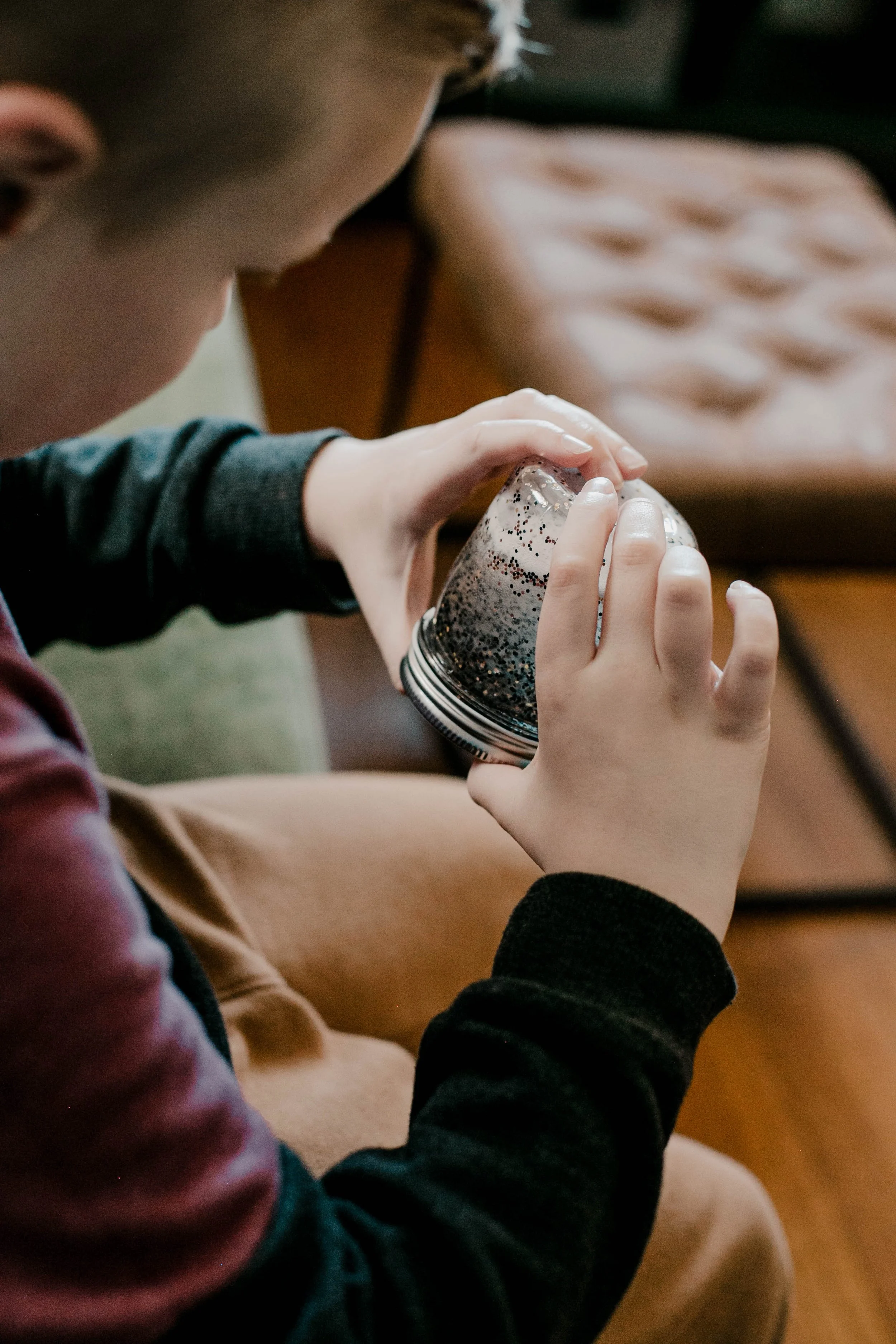 A person holding a jar filled with glitter or small shiny objects, sitting on a chair in a cozy indoor setting.