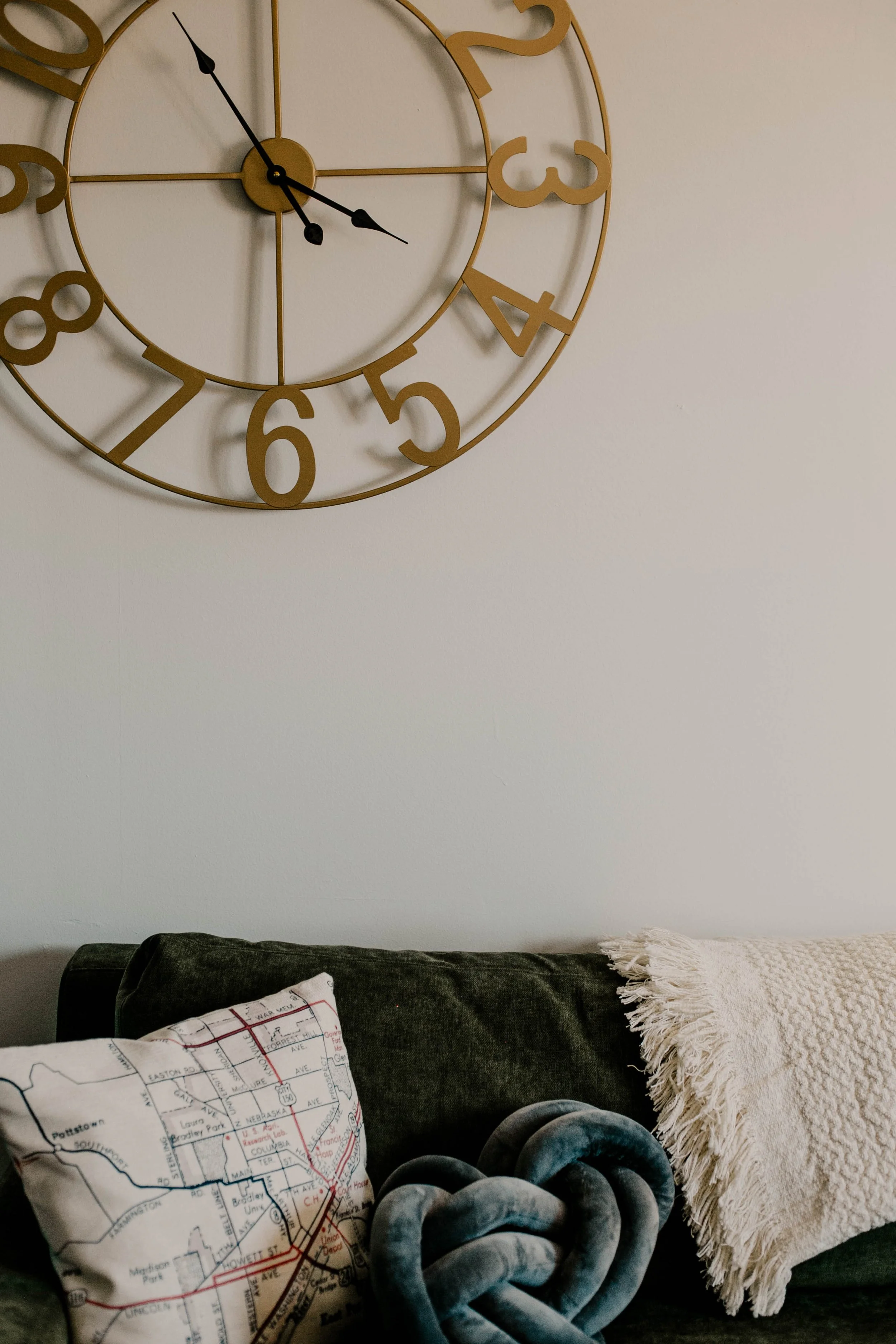 A wall clock with gold numbers and black hands, a pillow with a subway map design, a blue knot-shaped cushion, and a cream-colored fringed throw blanket on a dark couch.