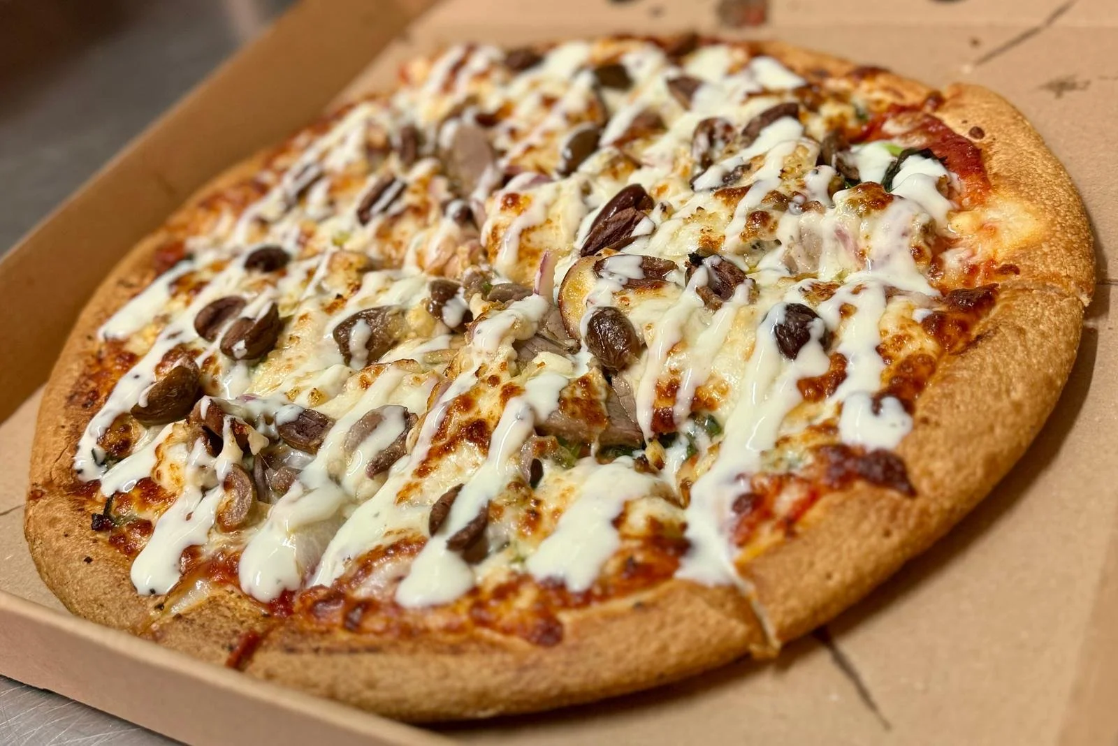 Jessie's Pizza A pizza with cheese, mushrooms, black olives, onions, and drizzled white sauce in a cardboard box. best pizza in Point Cook