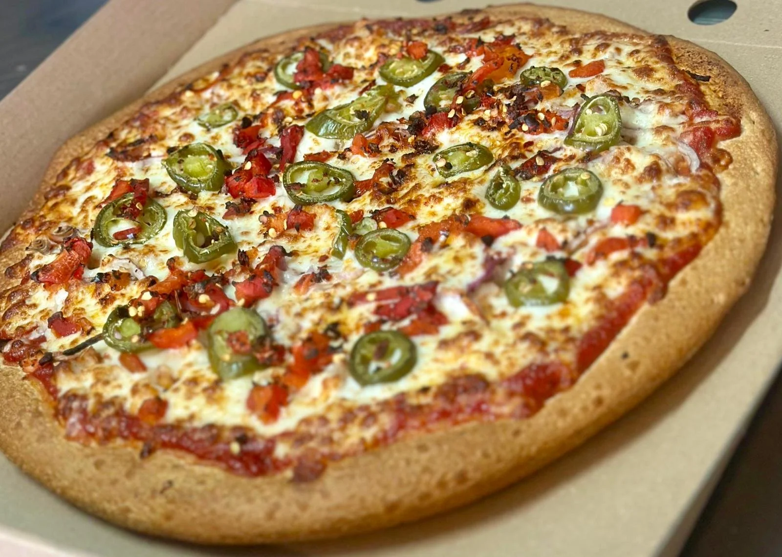 Jessie's Pizza A close-up of a large oval pizza with melted cheese, topped with sliced jalapeño peppers, diced tomatoes, and bits of bacon, inside a cardboard pizza box. best pizza in Point Cook