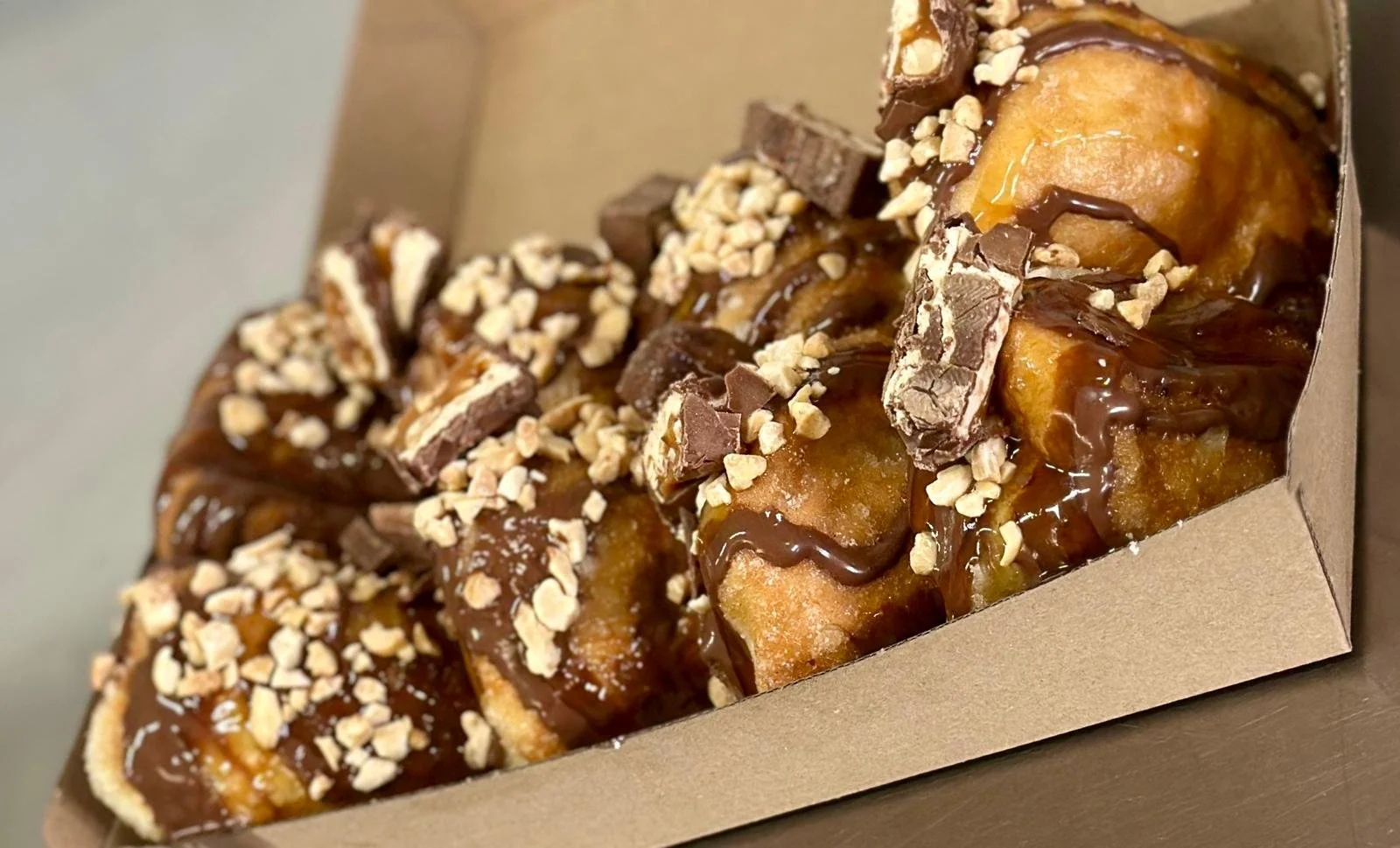 Jessie's Pizza A cardboard box containing several fried donuts topped with chocolate drizzle, chopped nuts, and pieces of chocolate candy. best pizza in Point Cook