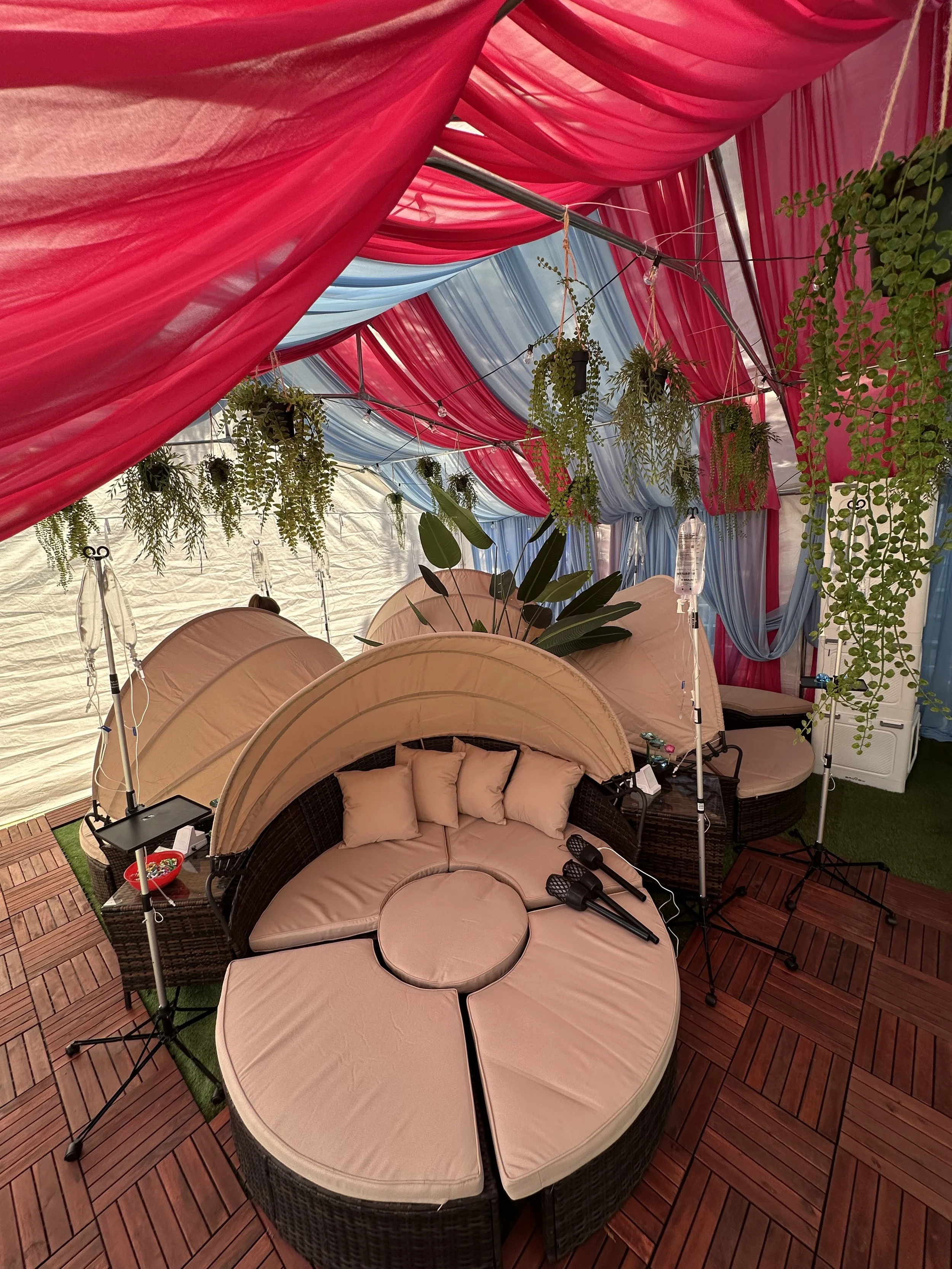 Colorful tent interior with pink and blue drapes, hanging plants, wicker seating arrangement with beige cushions, IV stands, and some small tables.