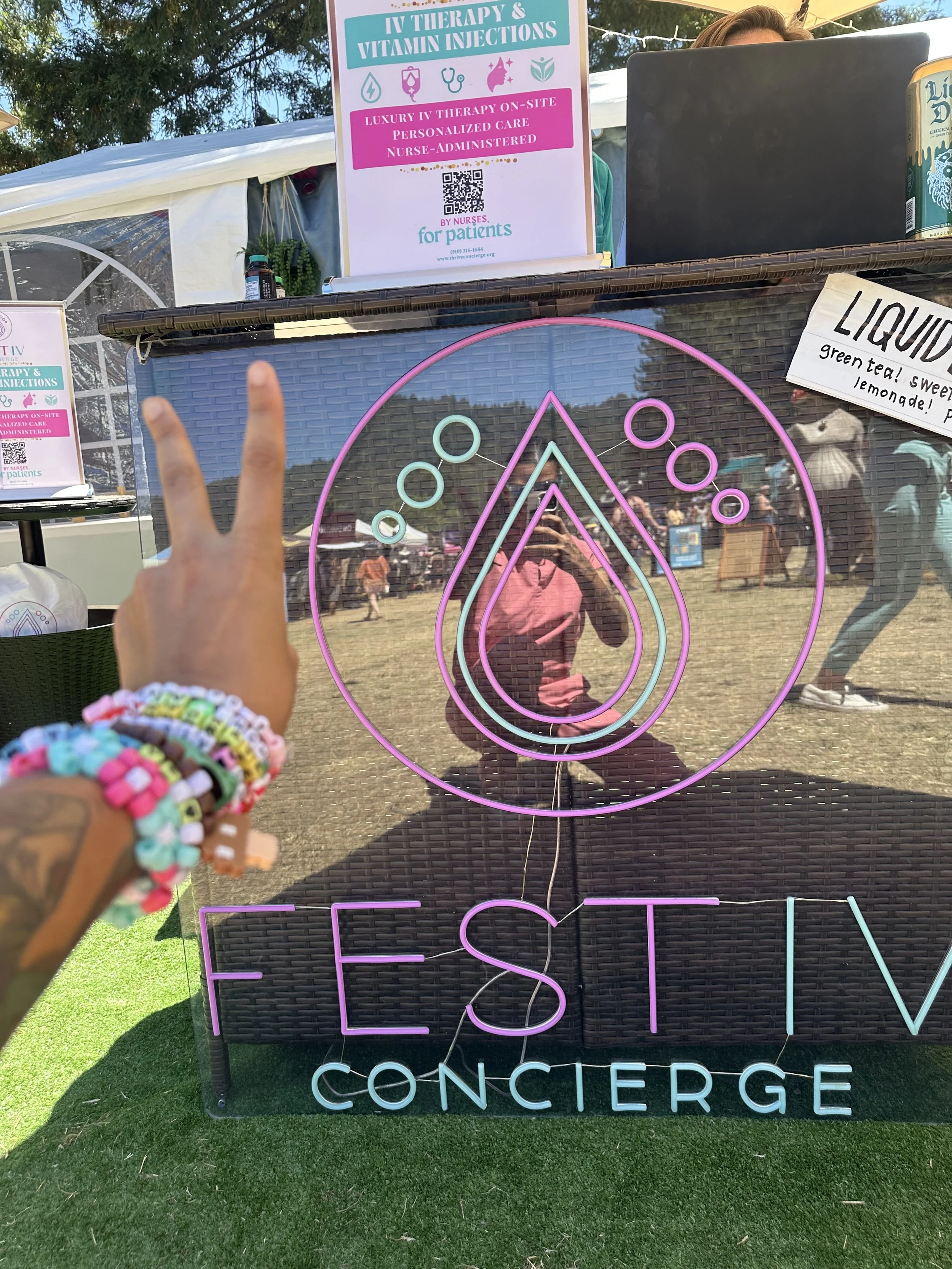 A person holding up a peace sign in front of a neon sign that says "FESTIV CONCIERGE." The person is wearing colorful beaded bracelets. The neon sign is reflected in a glass surface, showing a reflection of a person in a pink shirt taking the photo a