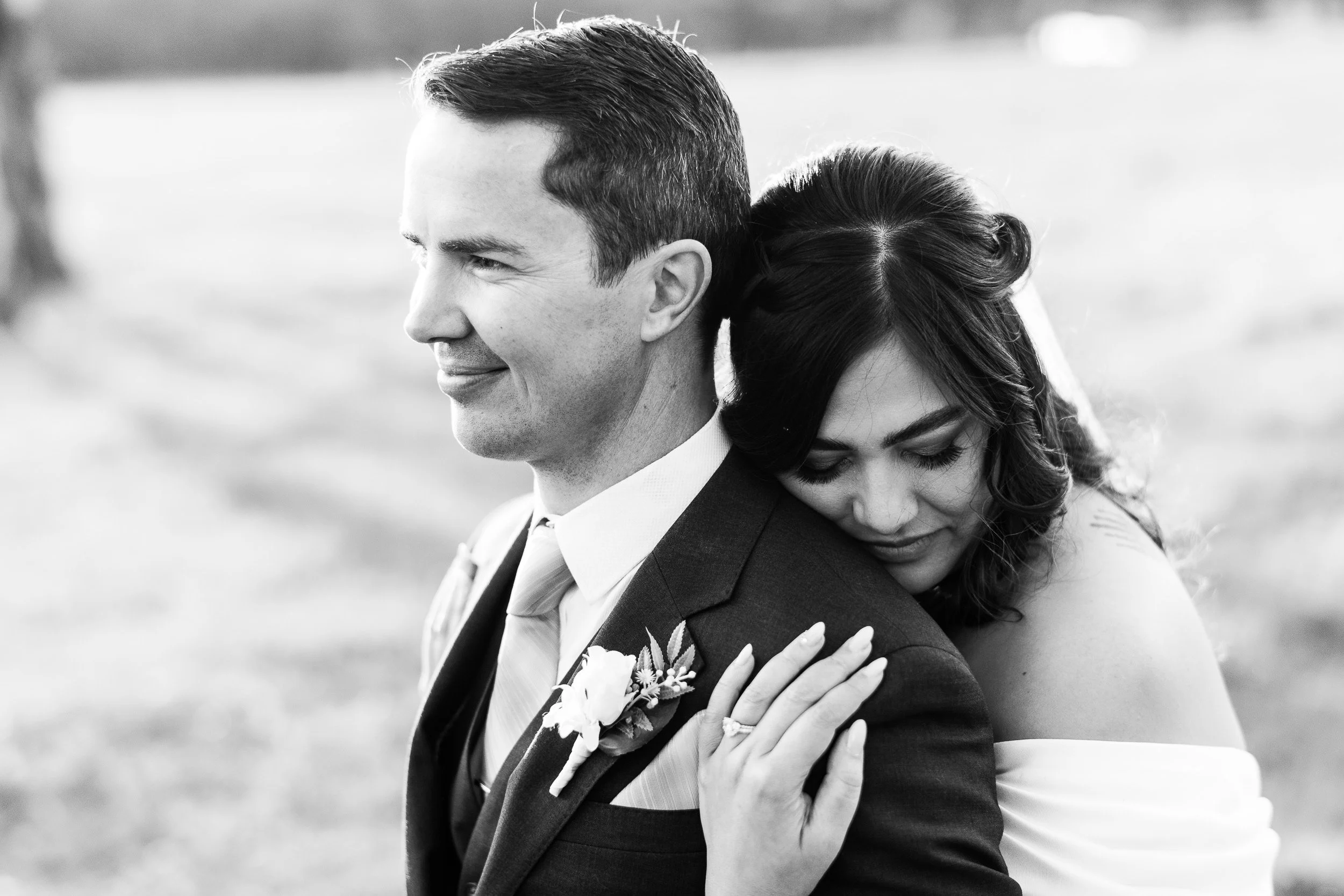 Black and white photo of a bride hugging a groom from behind, both smiling softly, outdoors, with a grassy background.