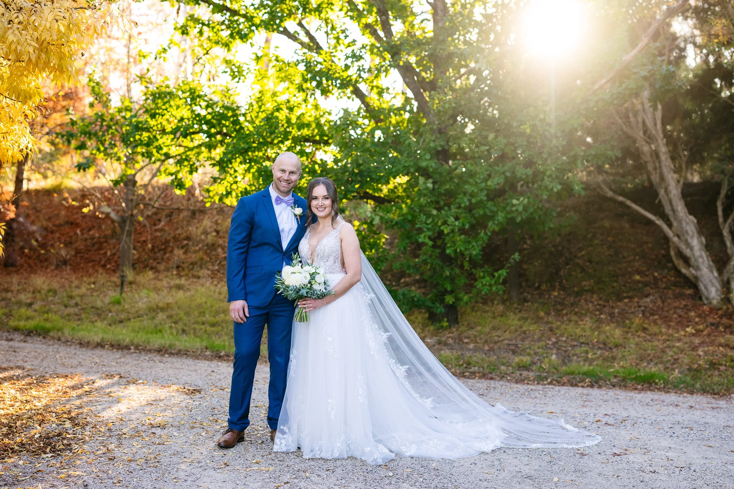 A bride and groom standing together outdoors during sunset, smiling. The bride is holding a bouquet of white flowers, wearing a white wedding gown with lace details and a long veil. The groom is dressed in a blue suit with a bow tie and boutonniere. Trees with green and autumn-colored leaves are in the background.