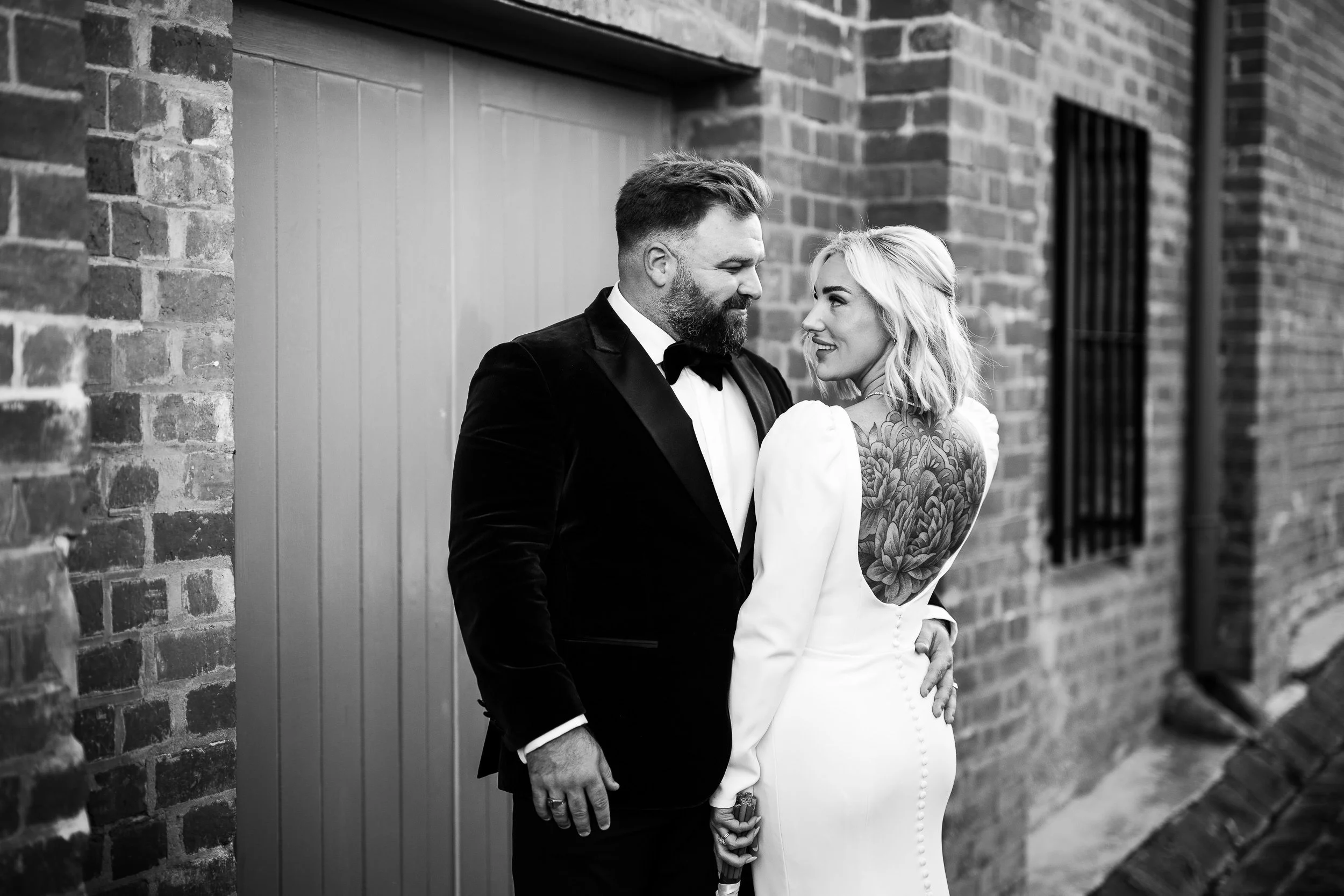 A black and white photo of a couple dressed in formal attire, standing close and looking into each other's eyes against a brick wall.