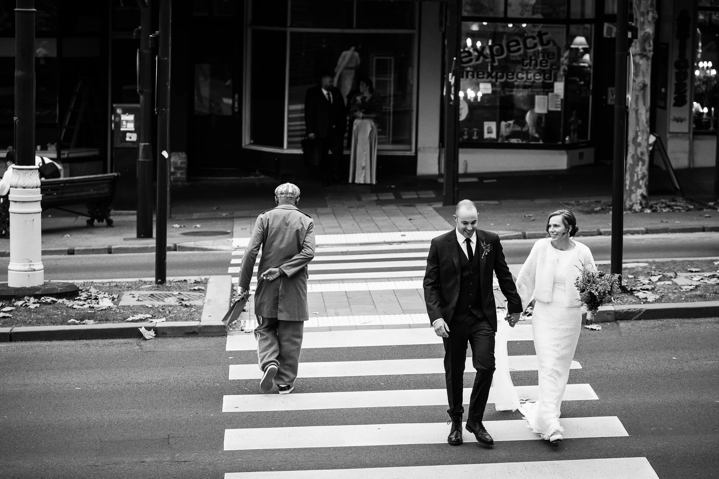 A couple dressed in wedding attire holding hands and walking across a crosswalk in an urban setting, with a man in a trench coat and hat crossing in the opposite direction behind them.