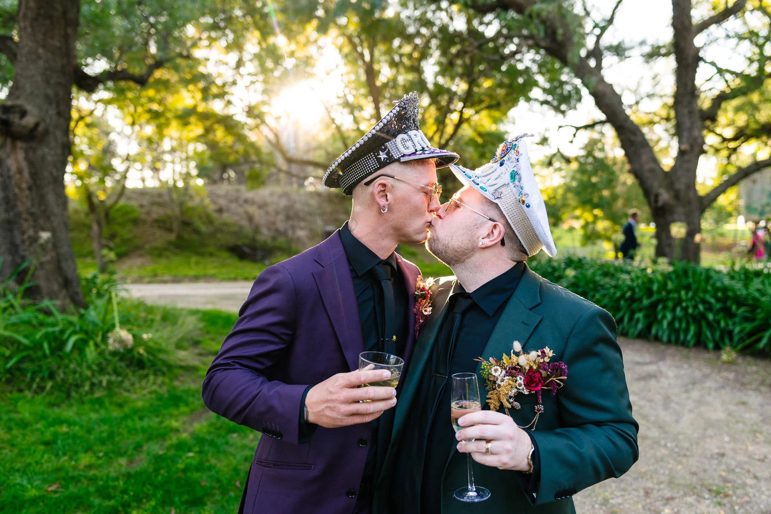 Two men in suits and decorated hats kissing outdoors during a celebration, each holding a glass of champagne, with trees and sunlight in the background.