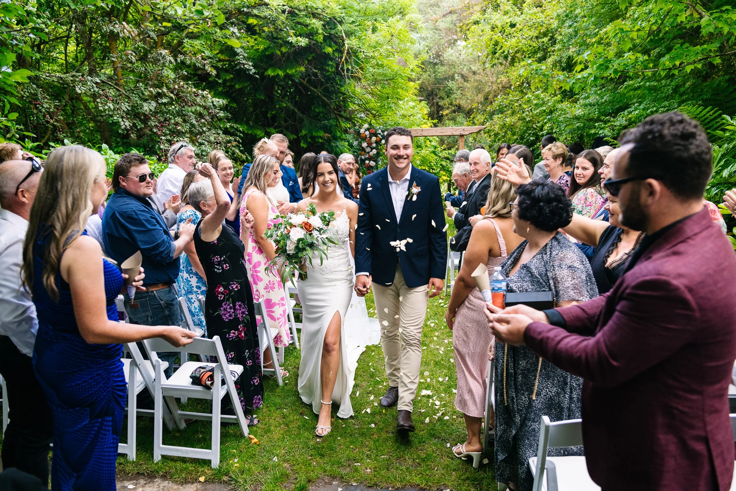 Bride and groom walking down the aisle at their outdoor wedding ceremony, surrounded by smiling guests in a lush green garden.