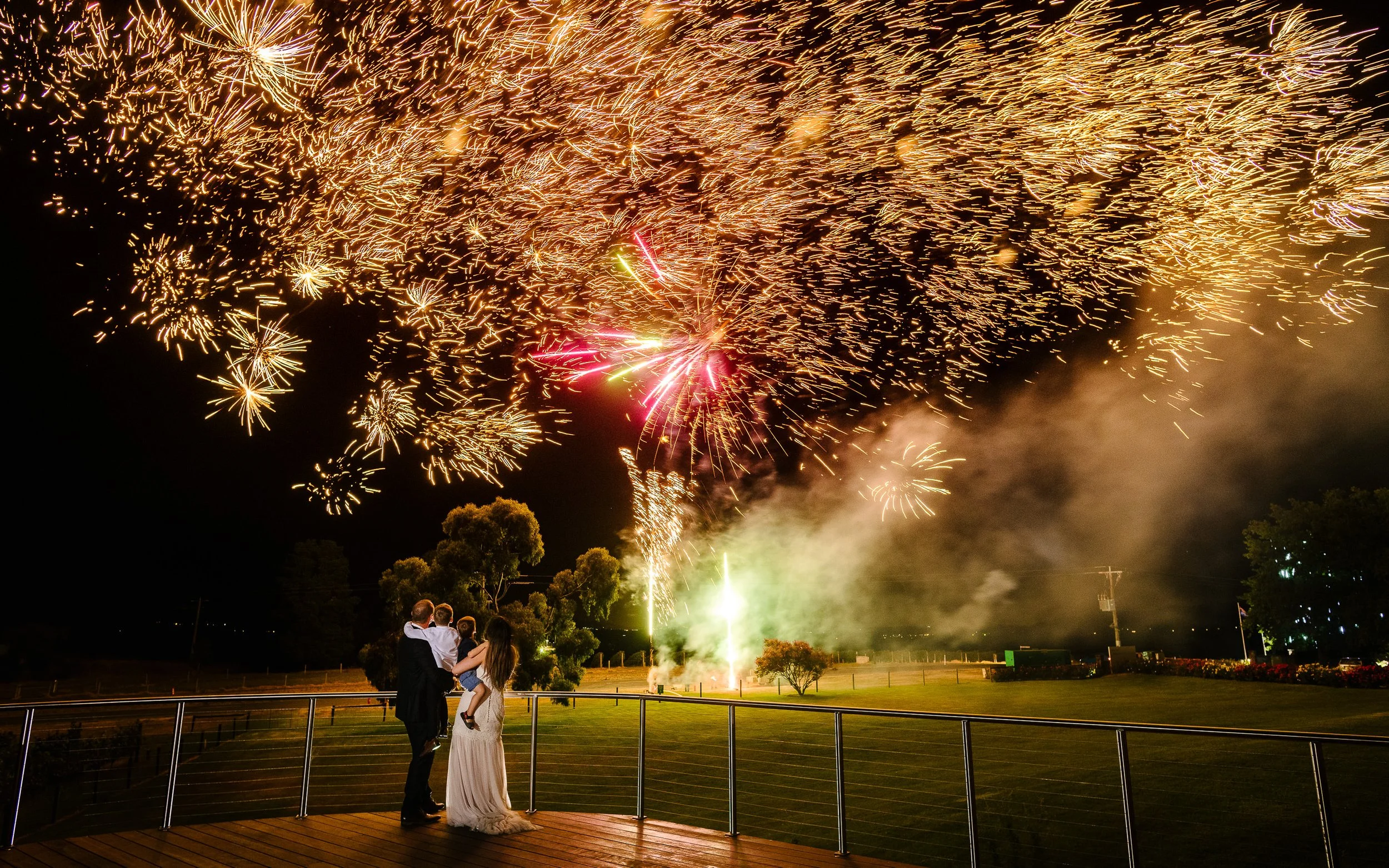 A family watching fireworks display at night on a balcony with grassy area, trees, and a dark sky.