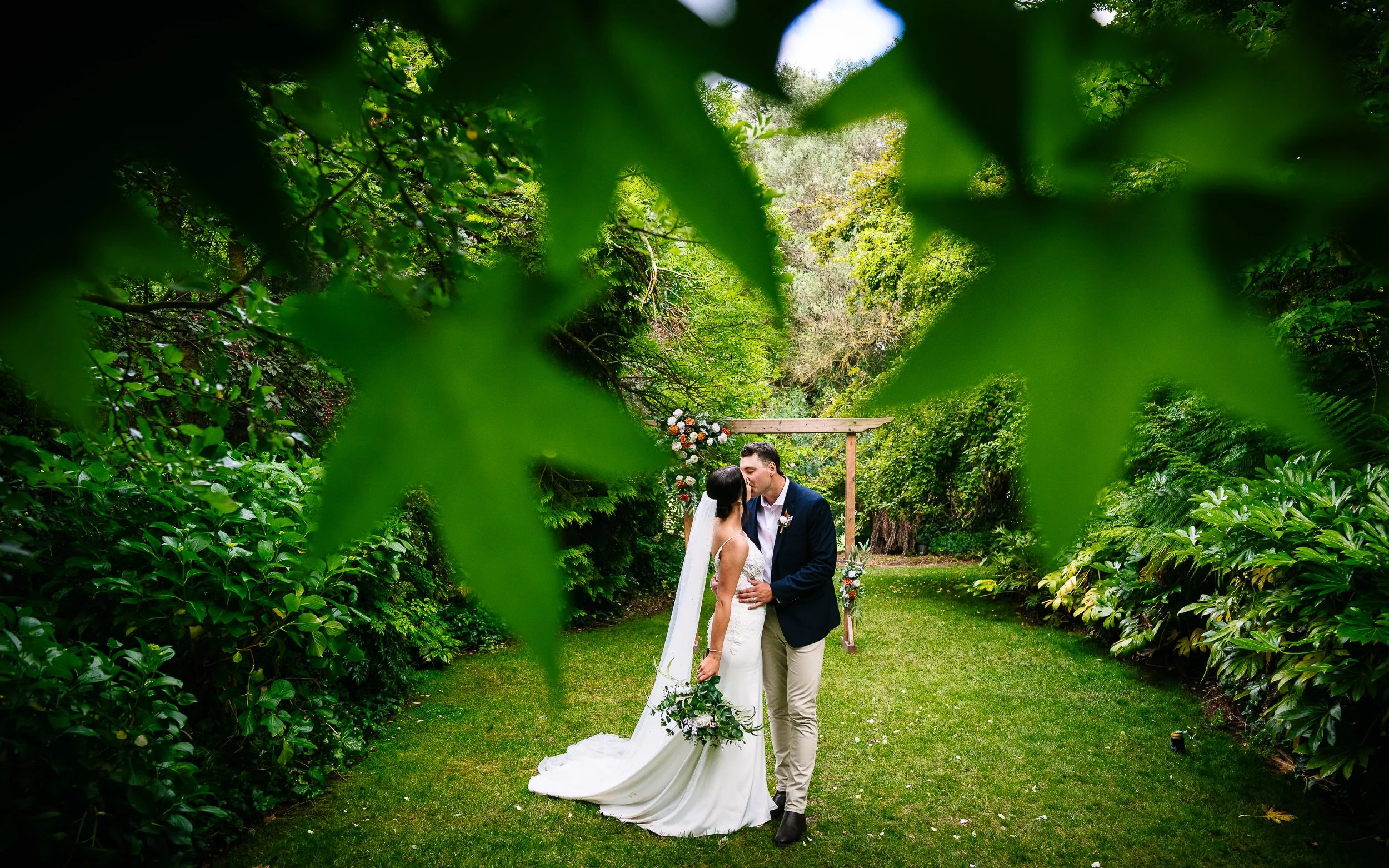 A bride and groom share a kiss during their wedding ceremony outdoors, framed by green foliage and a wooden arch decorated with flowers.