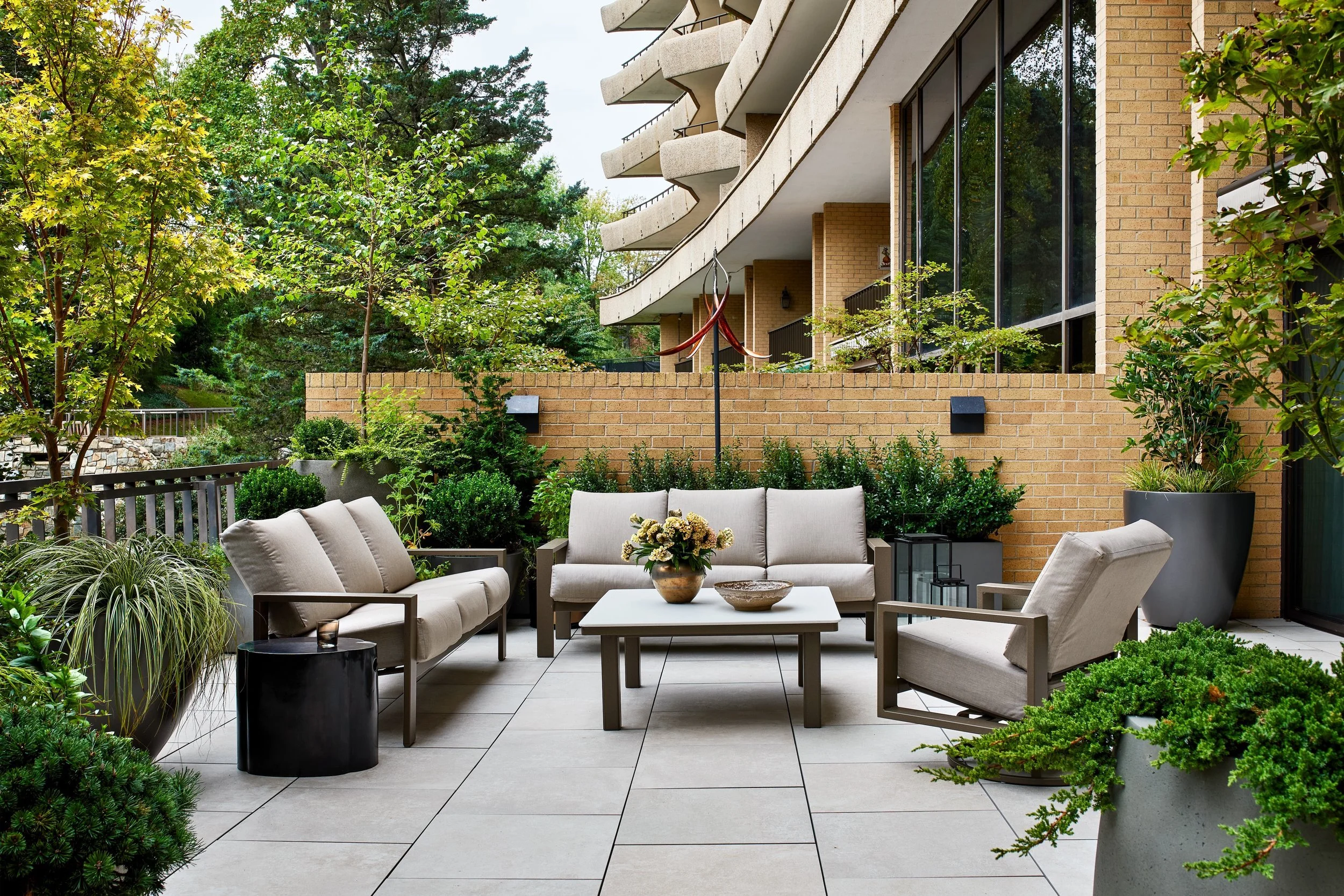 Contemporary terrace designed as an outdoor living room, featuring modern lounge seating, a low-profile table, and layered planters filled with greenery. Clean paving, integrated walls, and surrounding trees create privacy and a strong indoor–outdoor