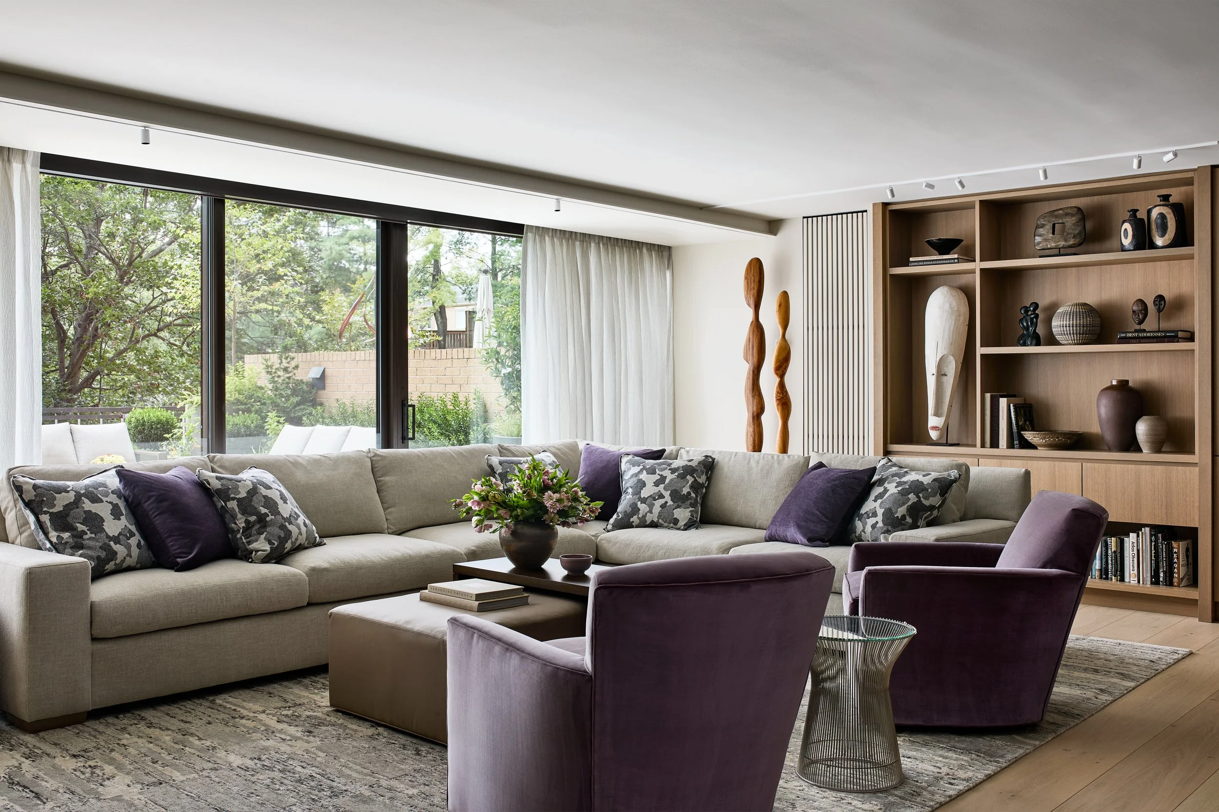 Serene contemporary living room featuring a custom sectional upholstered in soft neutral fabric, paired with sculptural plum lounge chairs and layered textiles. Floor-to-ceiling sliding glass doors frame lush treetop views, flooding the space with na