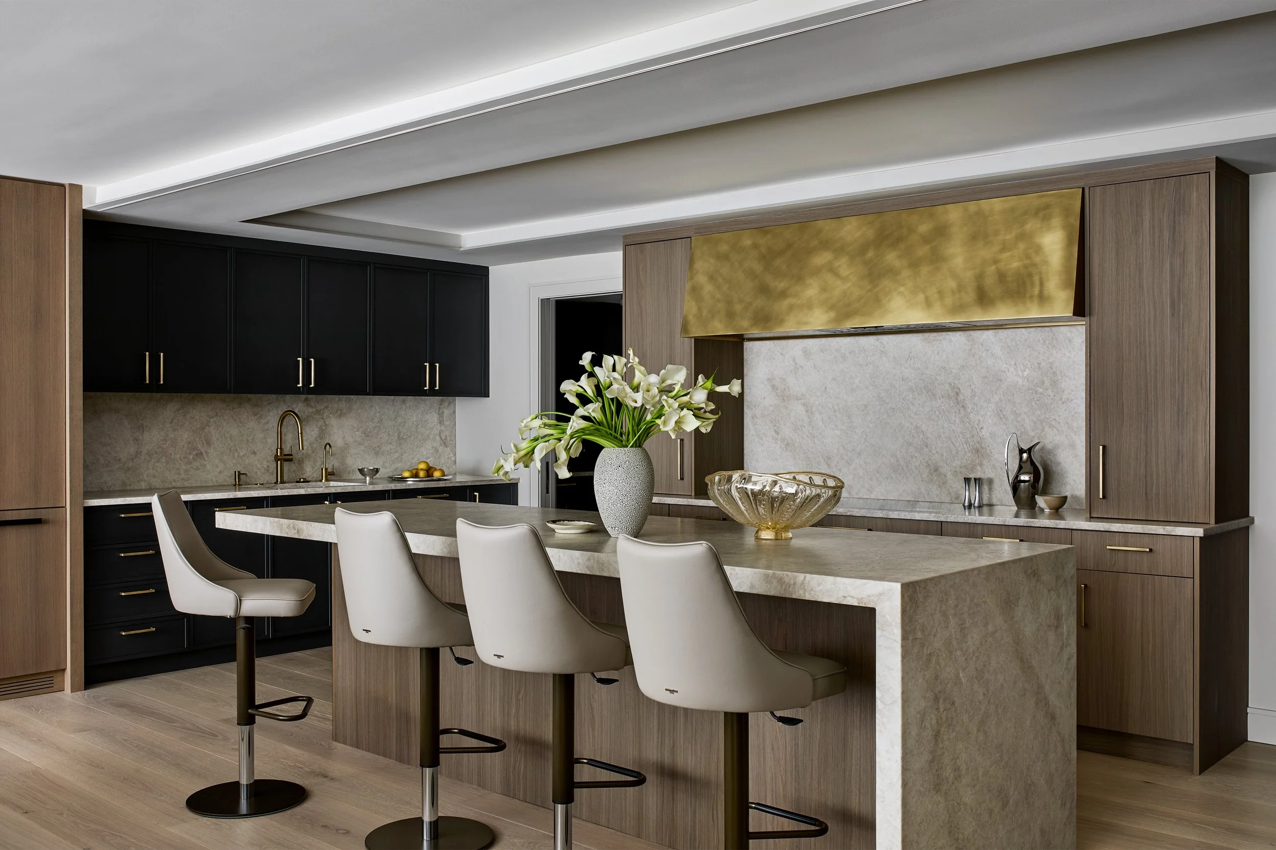Contemporary kitchen with a large stone waterfall island, sculptural brass vent hood, and custom cabinetry in warm wood and matte black finishes. Brass hardware and fixtures add warmth, while integrated storage and clean-lined millwork create a refin
