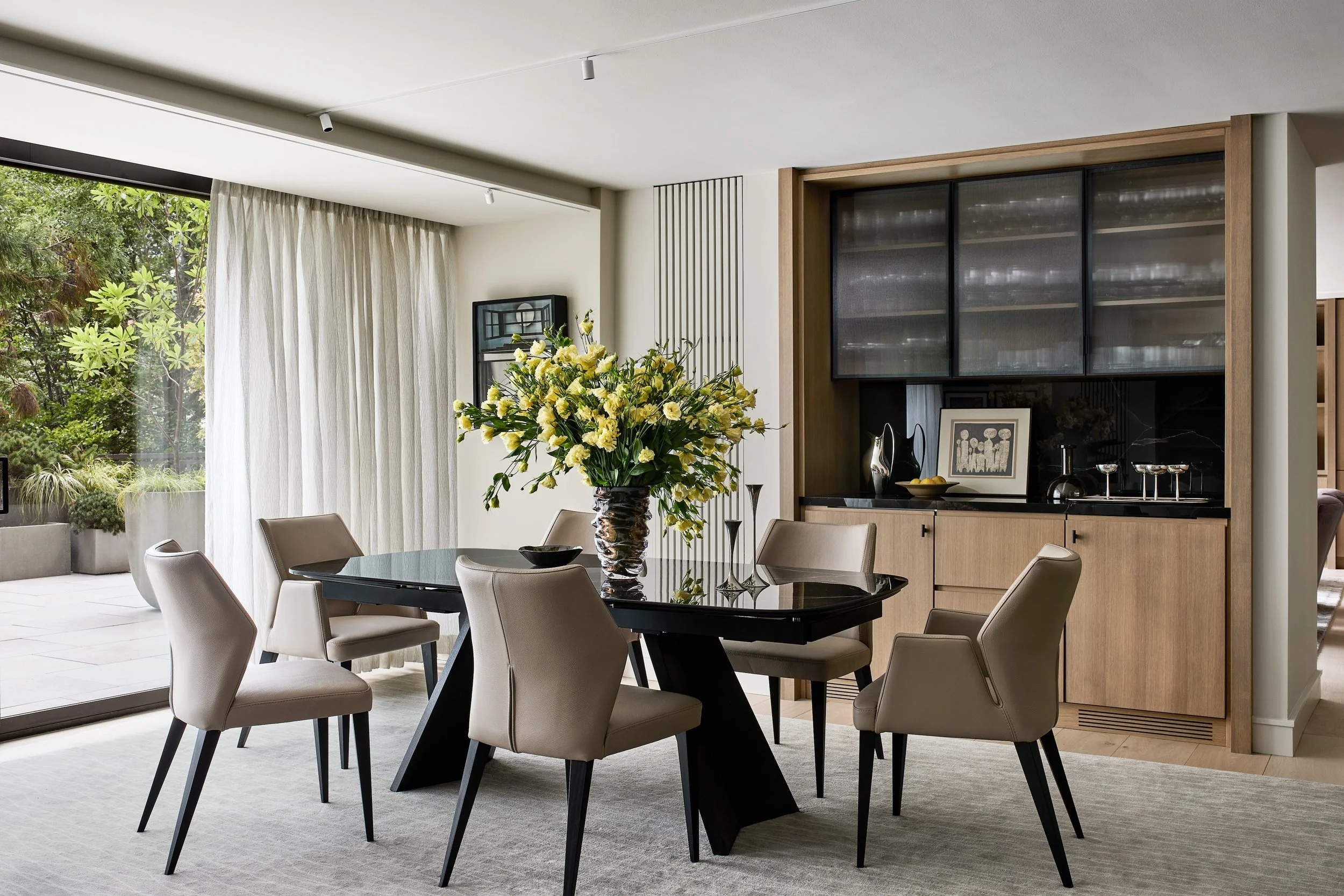 Modern dining room featuring a sculptural black dining table, upholstered chairs, and a custom oak built-in bar with ribbed glass cabinetry. Floor-to-ceiling windows and sheer drapery bring in natural light and views of surrounding greenery, while wa