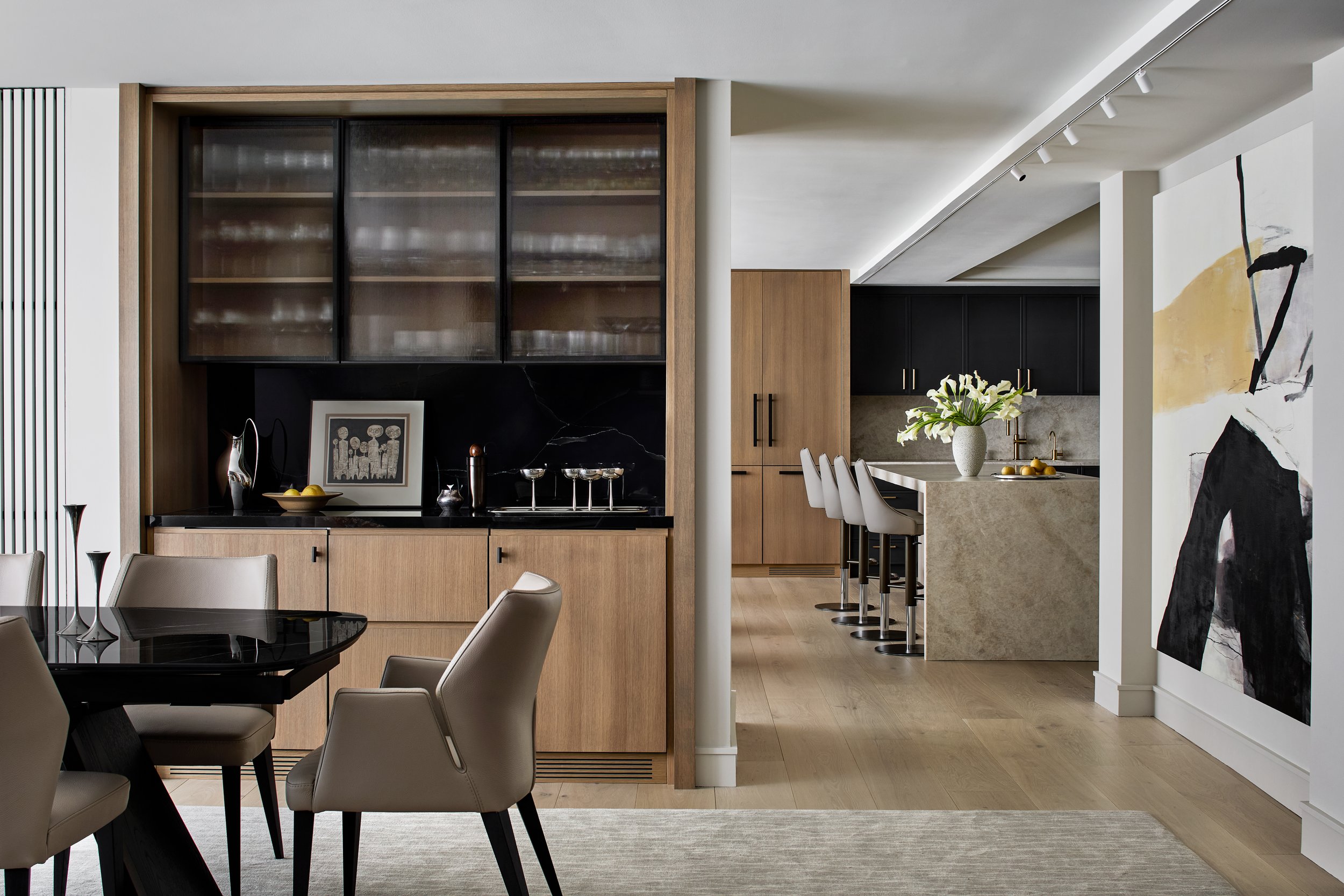 Open-plan dining and kitchen space featuring custom oak millwork, ribbed glass cabinetry, and integrated storage. A sculptural stone island anchors the kitchen, complemented by matte black cabinetry and brass hardware. Clean sightlines, layered light
