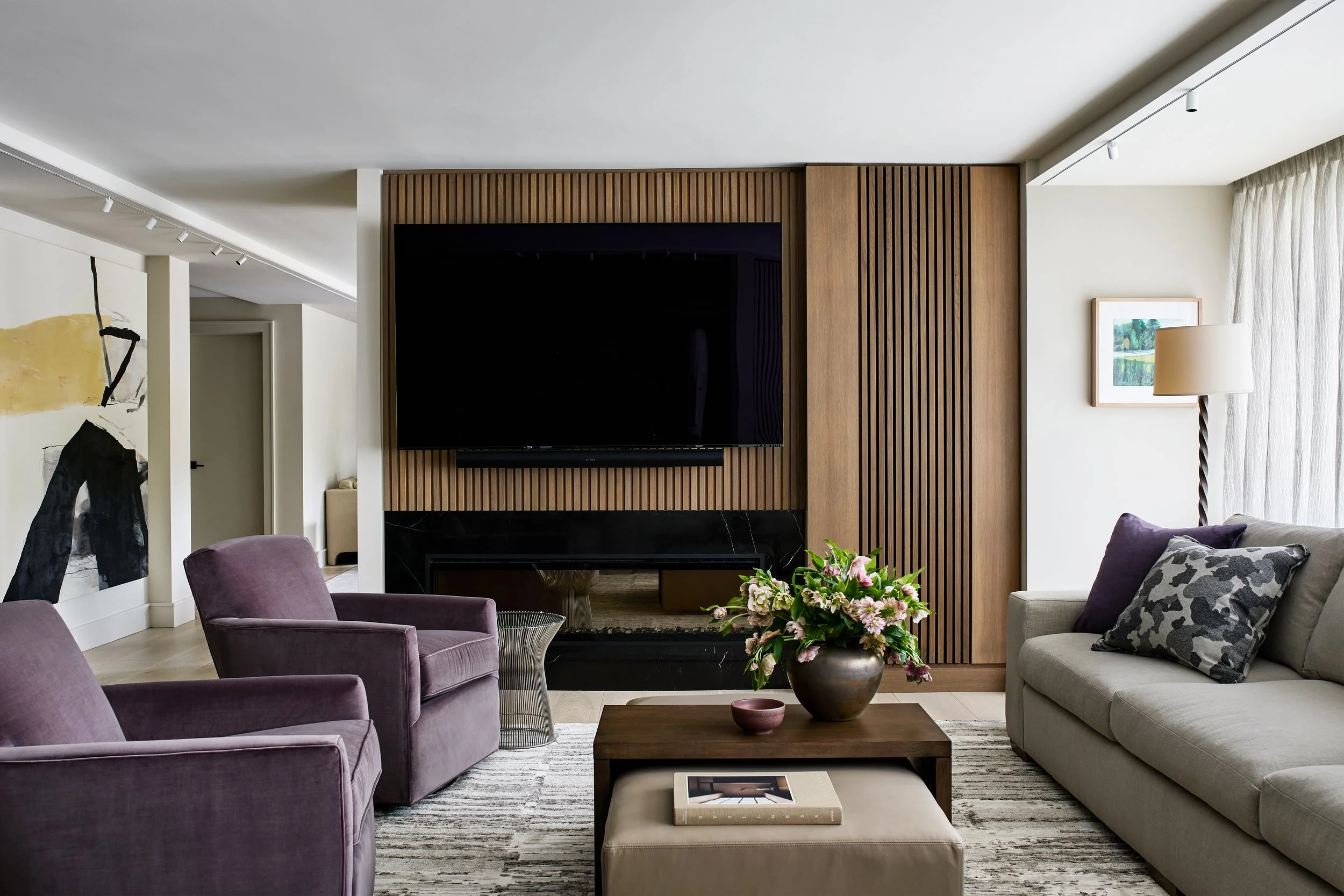 Contemporary living room with a custom slatted oak fireplace wall, integrated media center, and sculptural seating. Soft neutral upholstery is paired with plum lounge chairs, layered textiles, and curated art, creating a calm and inviting gathering s