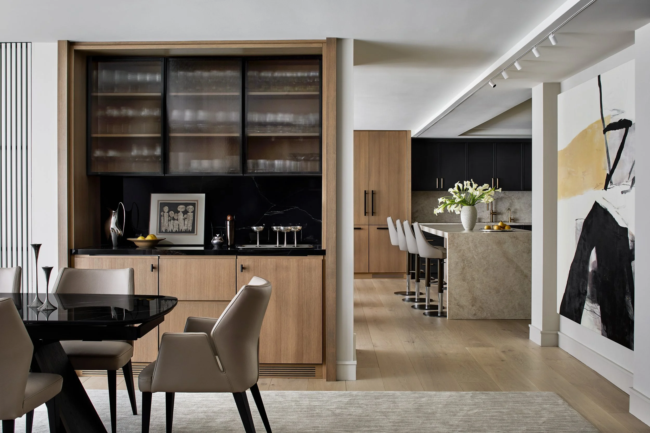 Open-plan dining and kitchen space featuring custom oak millwork, ribbed glass cabinetry, and integrated storage. A sculptural stone island anchors the kitchen, complemented by matte black cabinetry and brass hardware. Clean sightlines, layered light