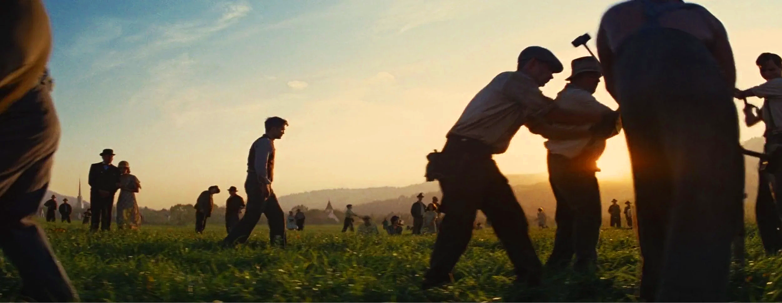 People working in a field at sunset, some are pulling on a rope, others are standing and observing, with a distant view of a church steeple and rolling hills.