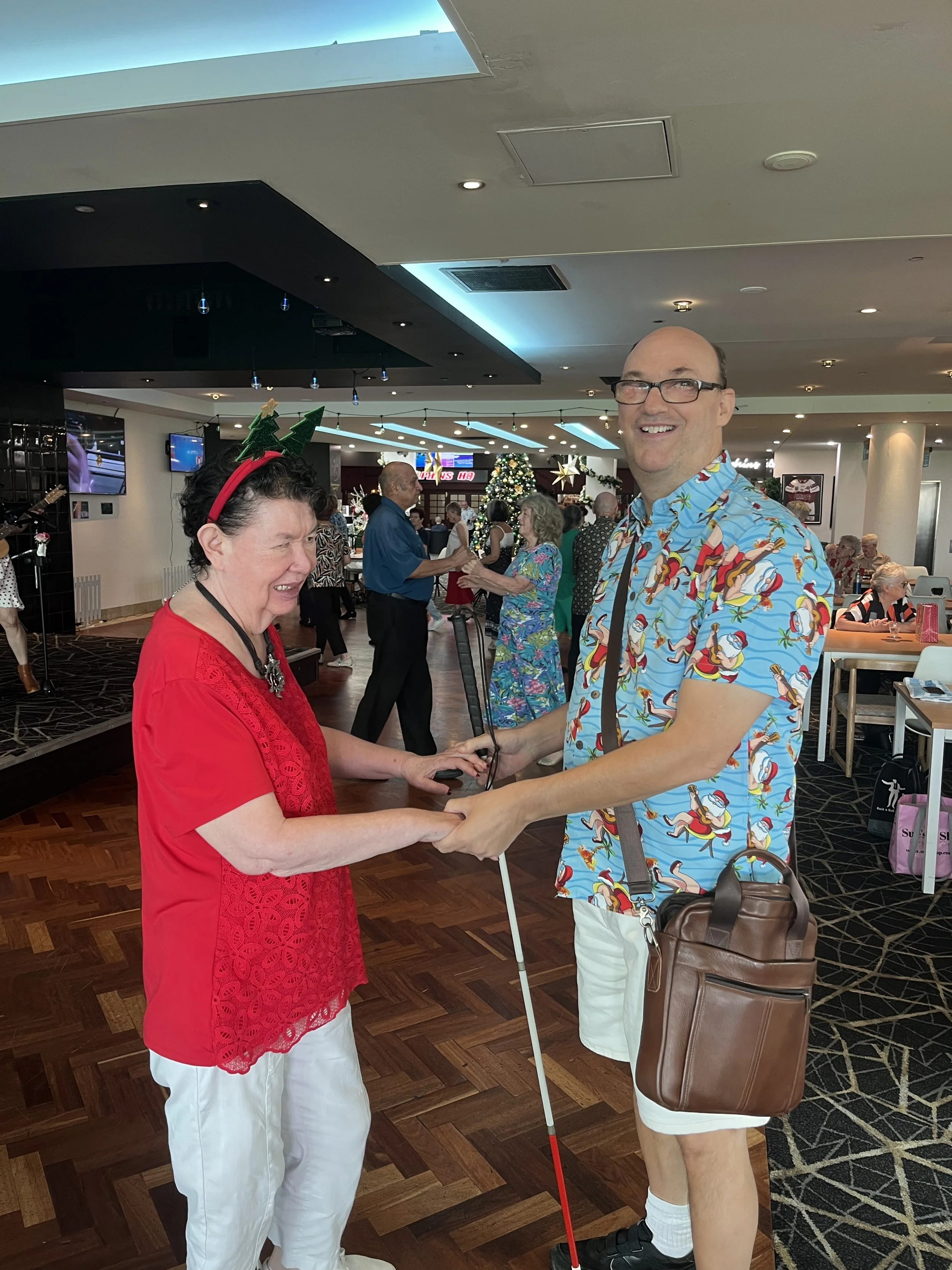 A joyful woman dressed in red with Christmas headbands and holding a cane is shaking hands with a tall man wearing a colorful holiday-themed shirt. They are at a holiday celebration in a decorated venue with other guests dancing and socializing in the background.