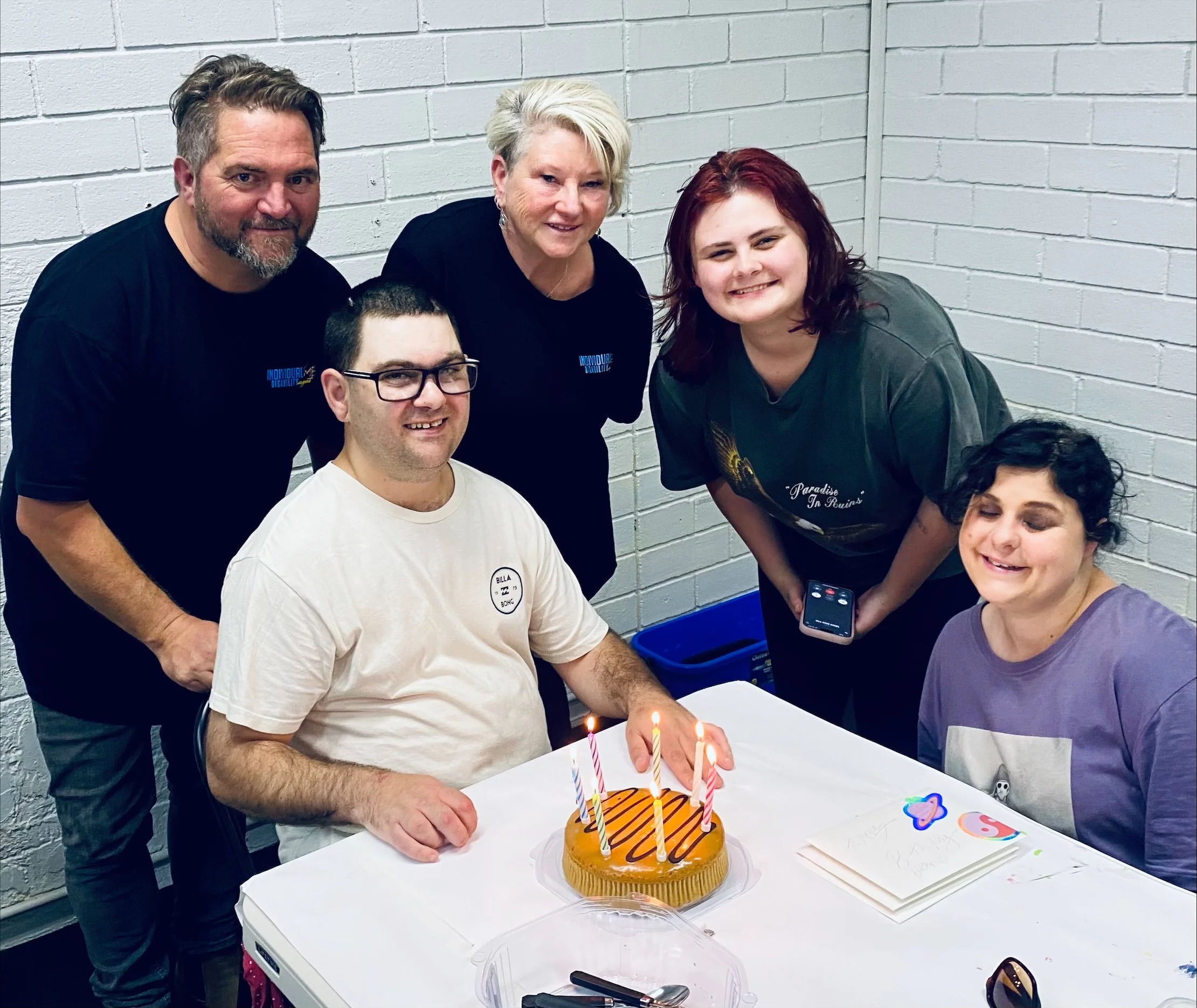 Group of six people celebrating a birthday with a caramel cake and lit candles at a table, in front of a white brick wall.