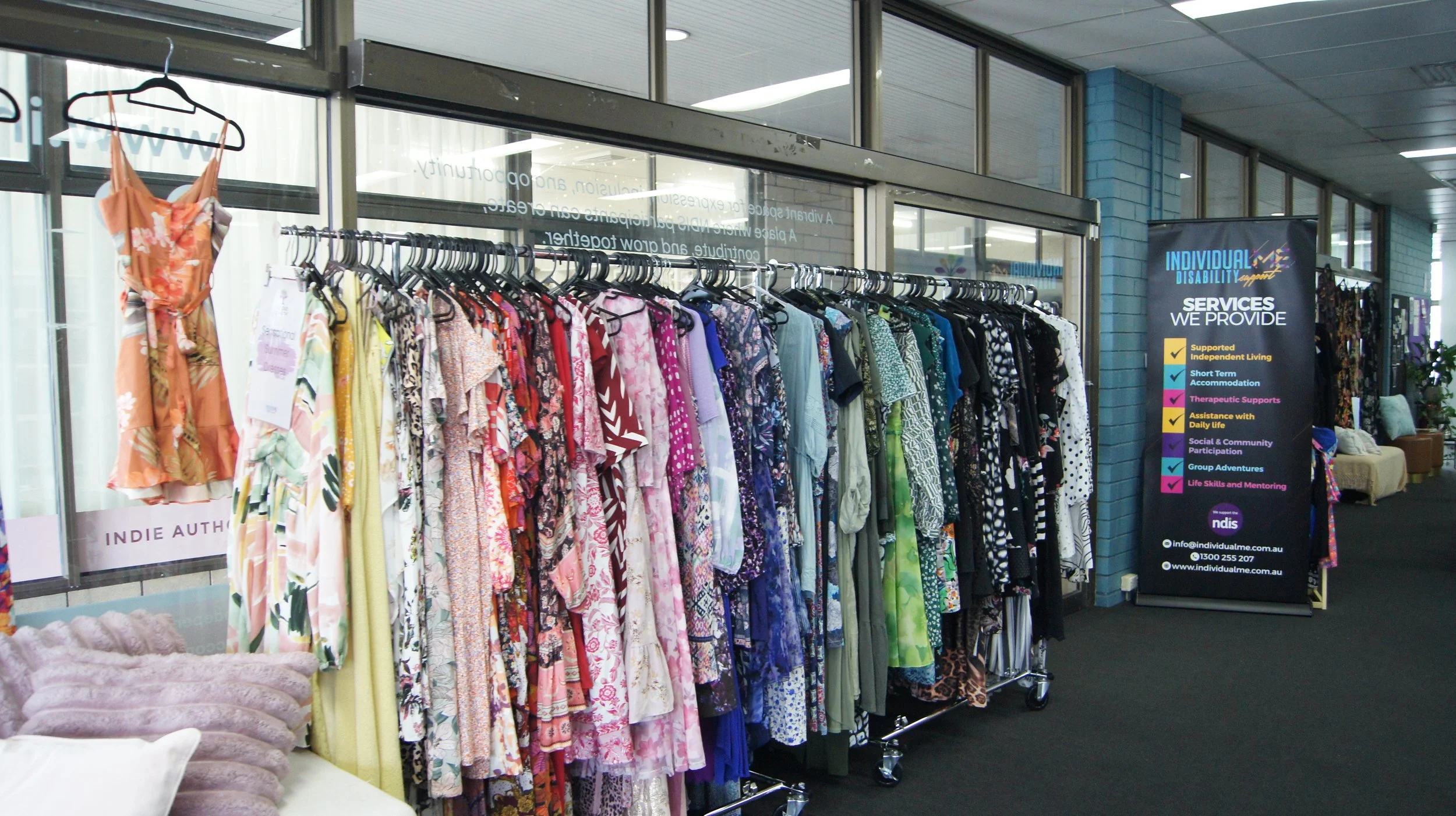 A clothing rack with colorful dresses and garments on hangers, situated inside an indoor space near a large window, with a sign listing services related to disability support and community participation.