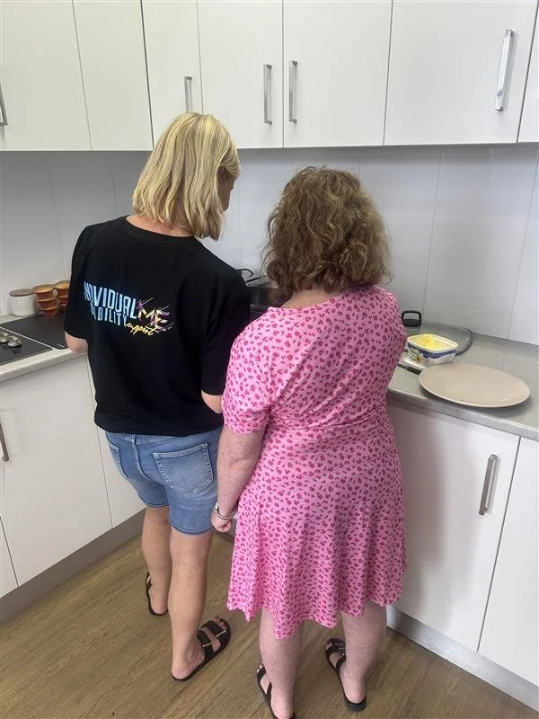 Two women are standing in a kitchen facing the counter, preparing food. One woman has blonde hair, is wearing a black t-shirt and denim shorts. The other has curly brown hair, is wearing a pink dress with purple patterns. There are kitchen cabinets above and a stove to the left.