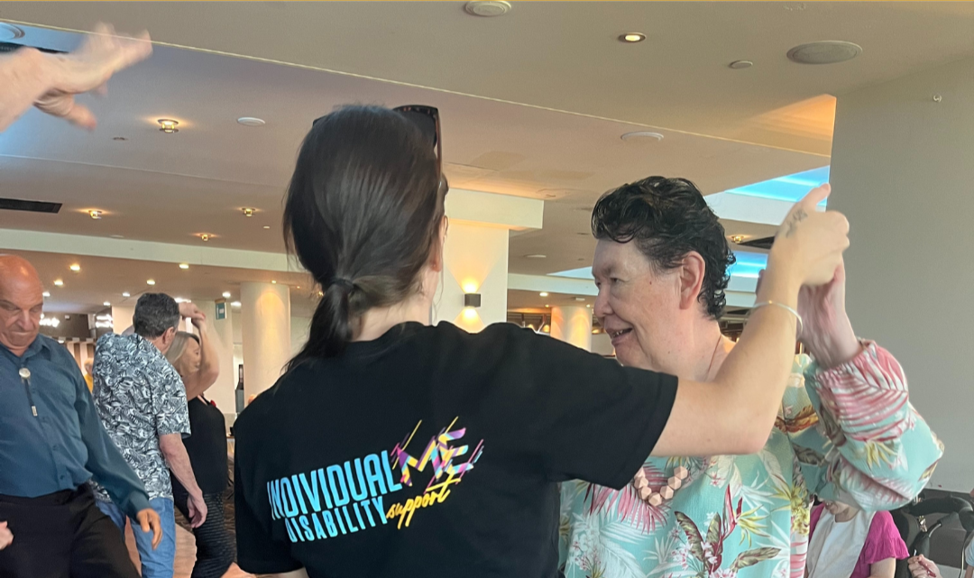 Two women dancing together in a social setting, with other people in the background. One woman is wearing a black t-shirt with text on the back, and the other is wearing a colorful, floral-patterned shirt.