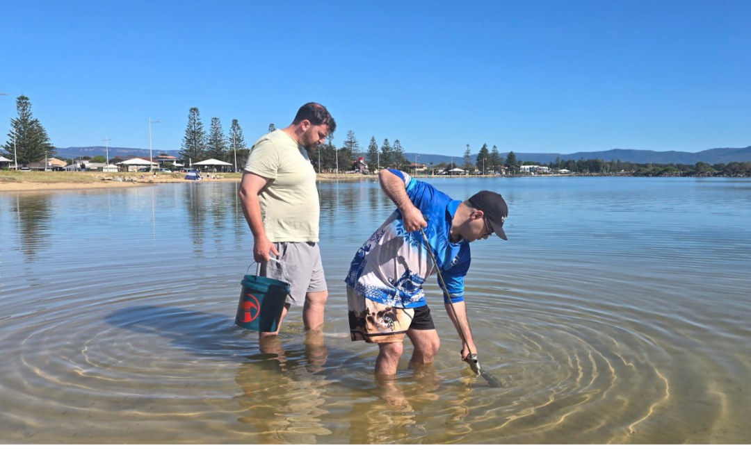 Two men standing in shallow water at a beach, one is holding a bucket and the other using a device to collect water or sediment, with houses and trees in the background under a clear blue sky.