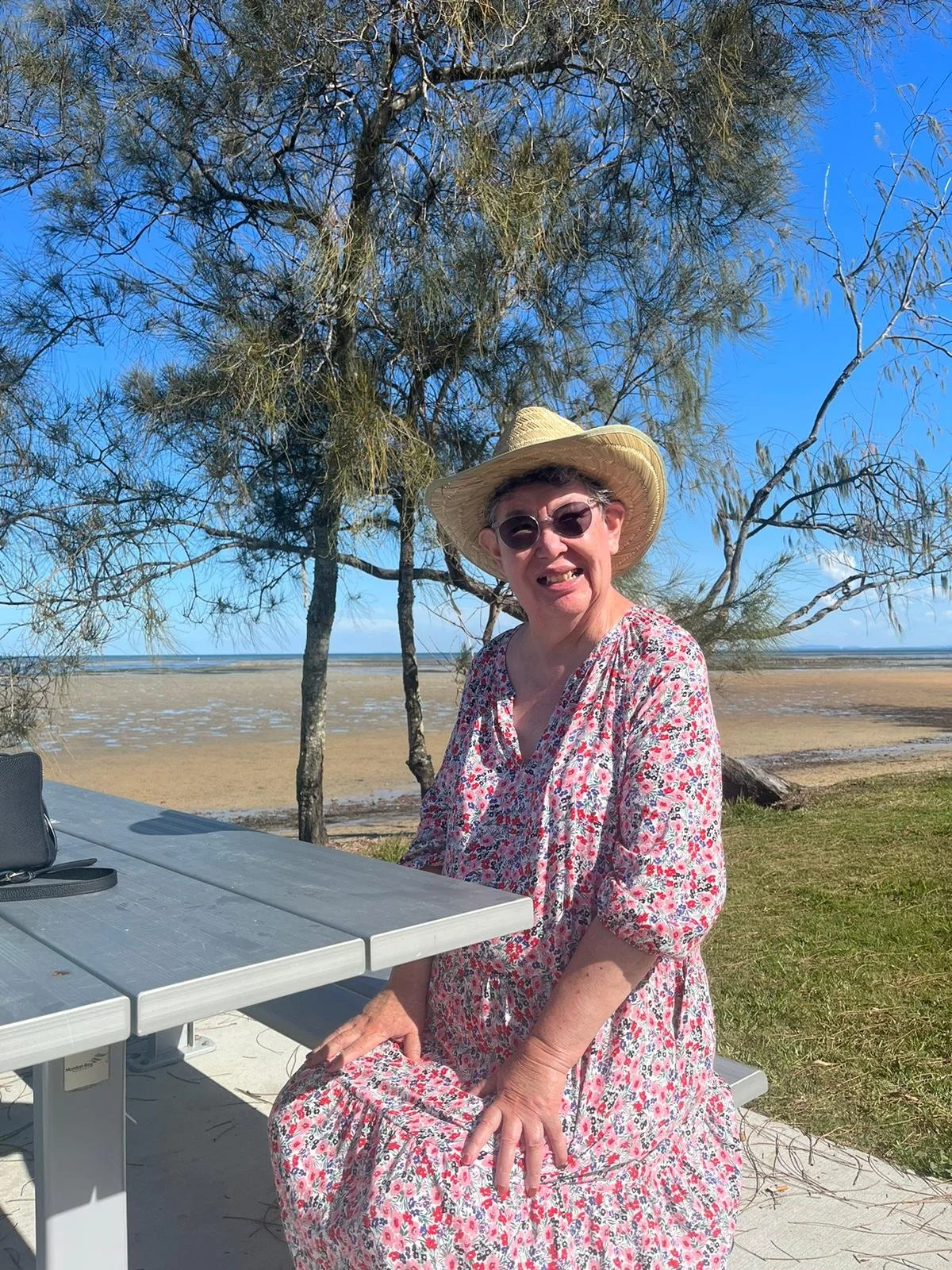 A woman sitting outdoors at a picnic table on a beach, wearing a floral dress, a straw hat, and sunglasses, making a playful face with her tongue out, with the ocean, sand, trees, and a blue sky in the background.