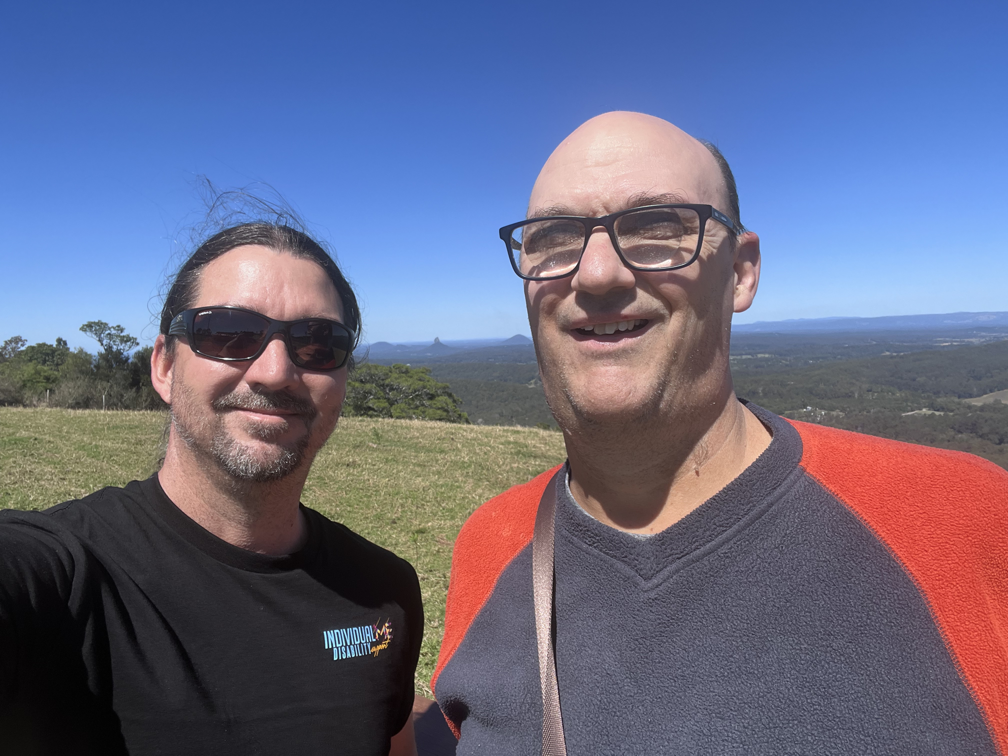 Two men smiling outdoors on a clear day, with a vast landscape and blue sky in the background.