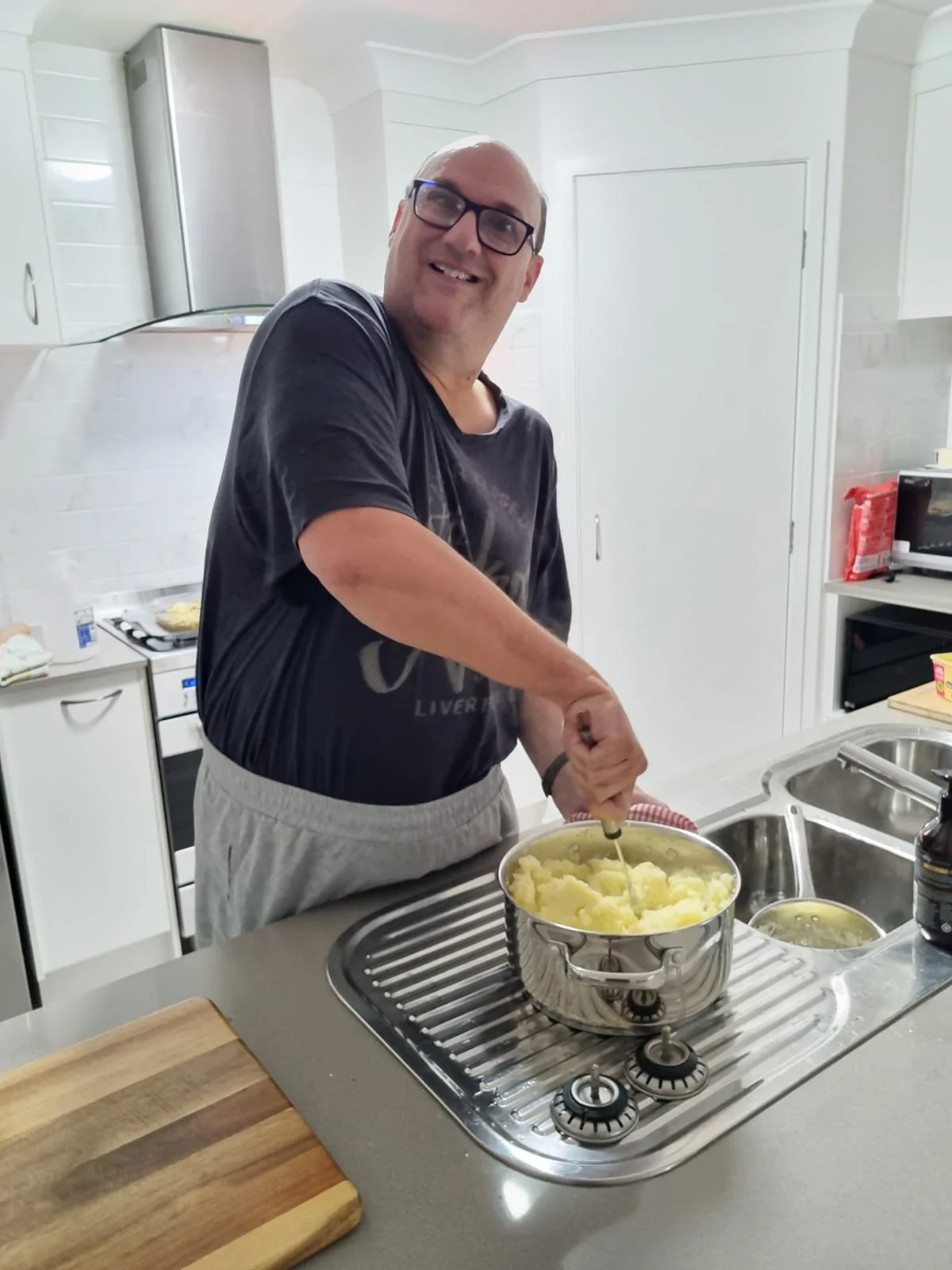 A man with glasses smiling as he stirs mashed potatoes in a pot on a kitchen stove.