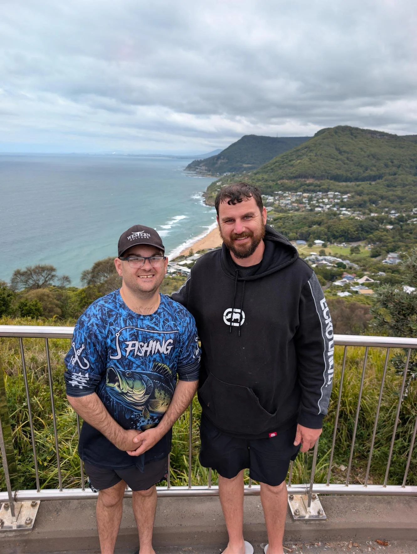 Two smiling men standing on a viewing platform overlooking a scenic coastline with ocean, beach, hills, and houses in the background.