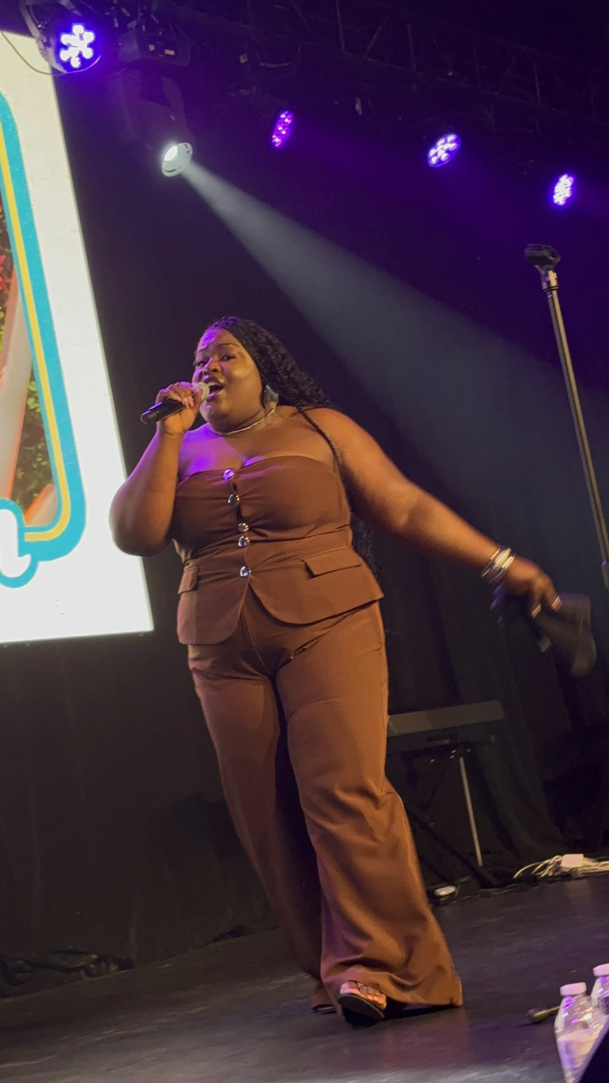 A woman singing on stage with purple stage lights and a large screen behind her, holding a microphone in her right hand.