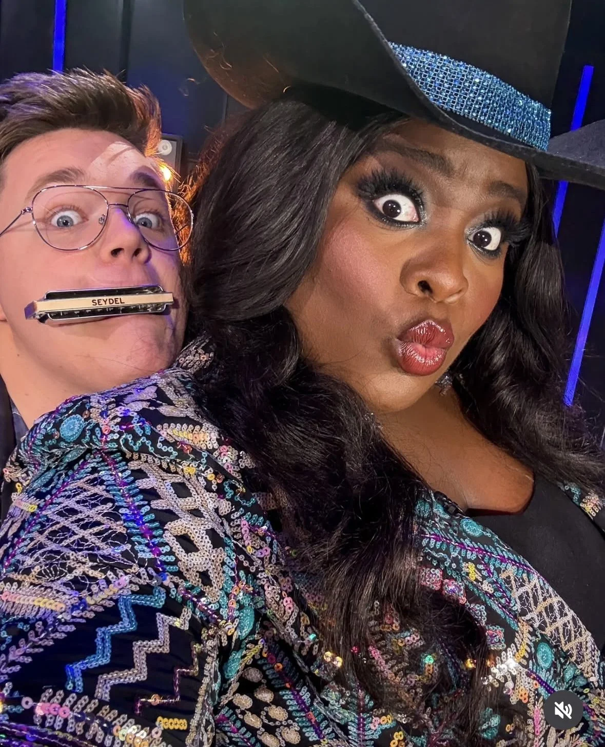 A woman wearing a decorative hat and a colorful, sequined jacket takes a selfie, with a man behind her holding a harmonica in his mouth. The woman has dark, wavy hair and dramatic makeup.
