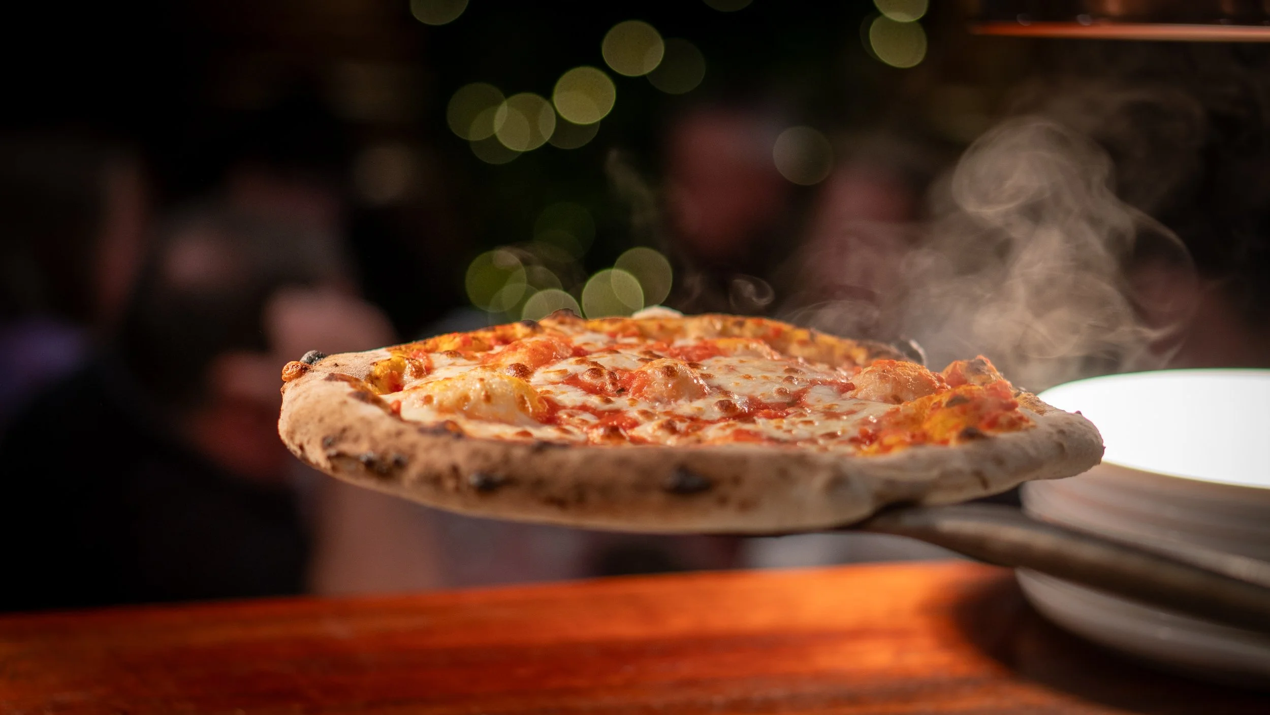Wood-fired margherita pizza on a wooden paddle with a blurred background.