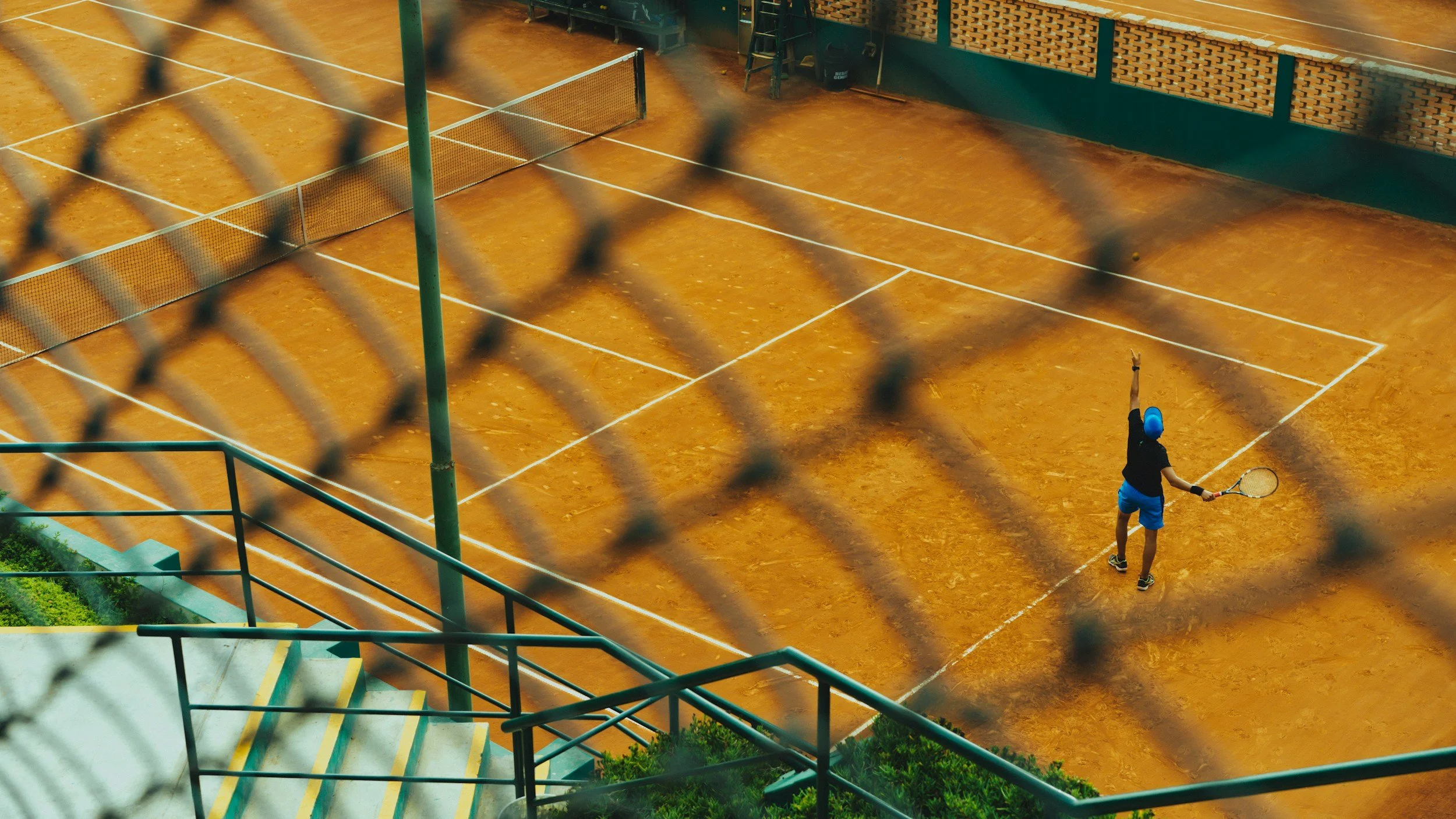 A person playing tennis on an orange clay court, seen through a chain-link fence