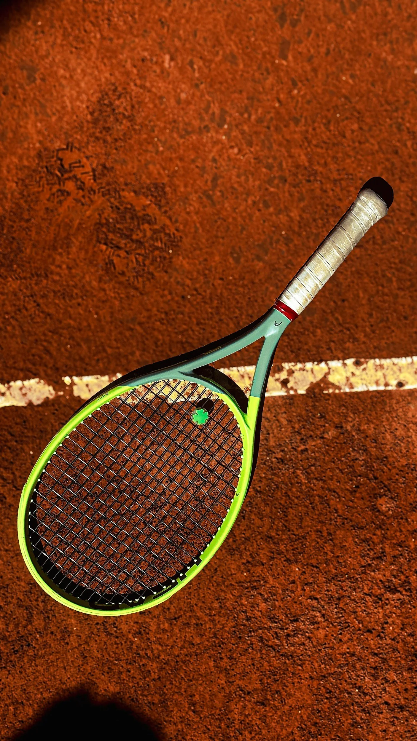 A tennis racket with a green and black frame and a four-leaf clover sticker on the strings, resting on a red clay tennis court with a white line.