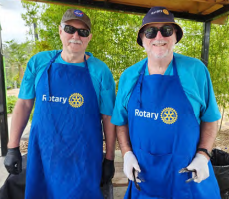 Two men wearing blue Rotary aprons and hats, sunglasses, and gloves, standing outdoors under a canopy, smiling at the camera.