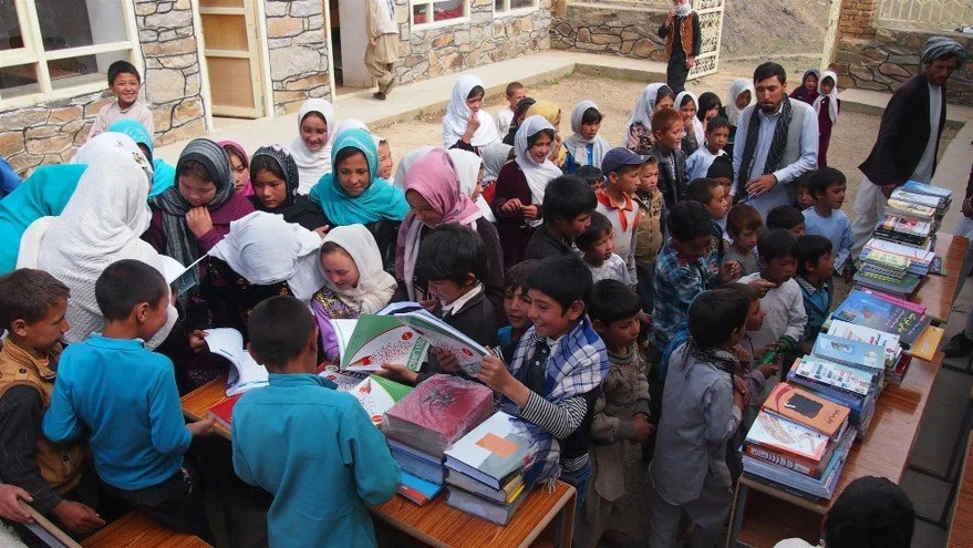 Children and adults gathered around tables with stacks of books at an outdoor event.