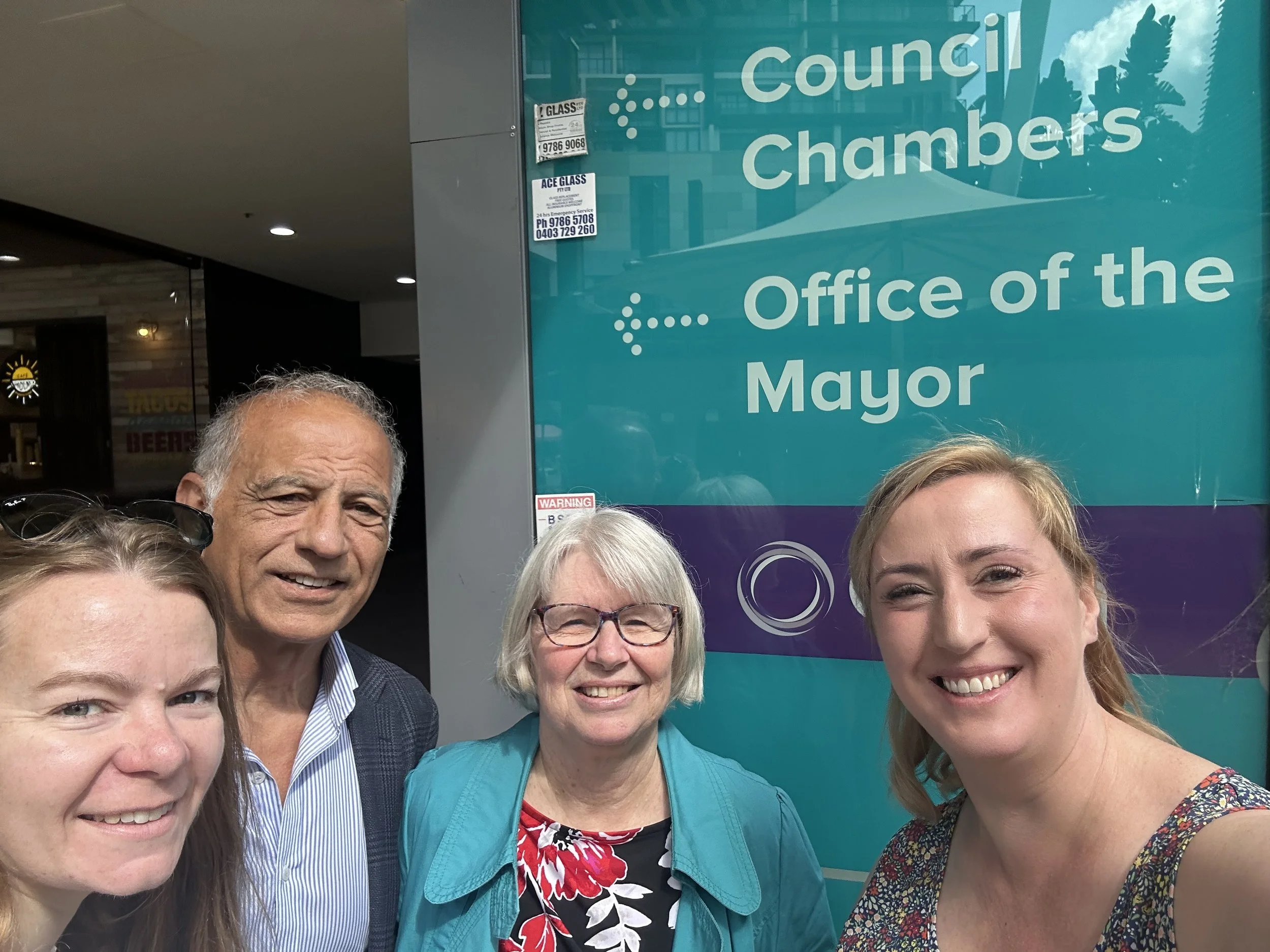 Four people smiling for a selfie in front of a sign that reads "Council Chambers Office of the Mayor."