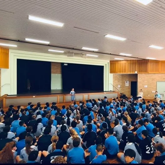 A large school auditorium filled with students sitting on the floor and facing a stage where a person is speaking at a podium.