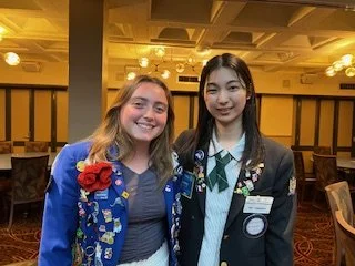 Two young women smiling at the camera in a restaurant or banquet hall setting, dressed in formal and semi-formal attire.